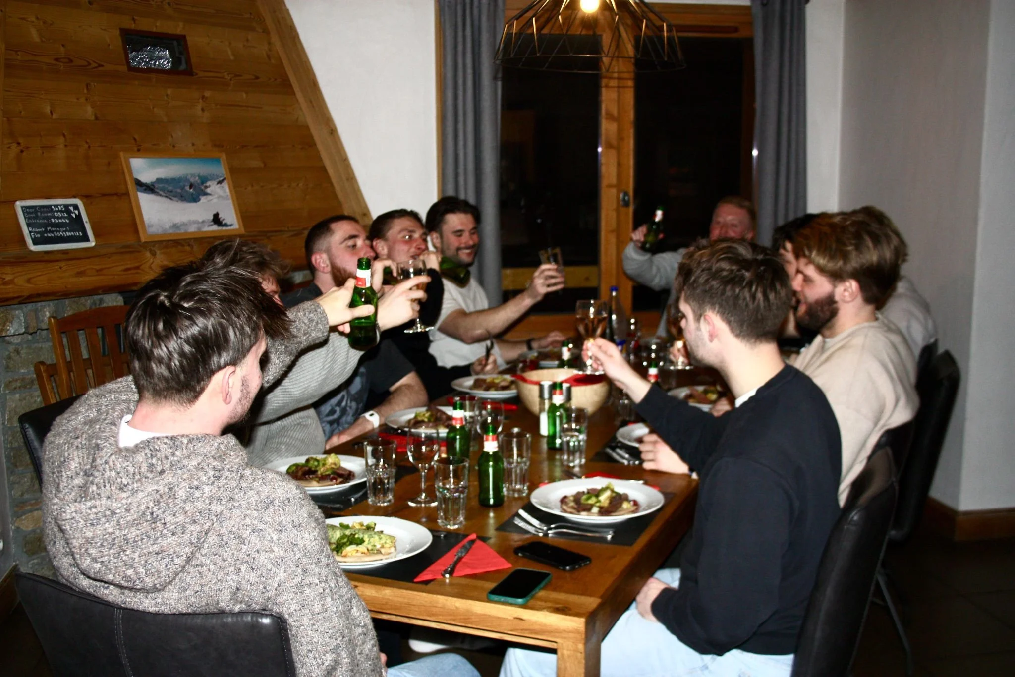 Group of people gathered at a wooden dining table, raising glasses in a toast, with plates of food and bottles of beer or soda in front of them, in a rustic indoor setting with wood-paneled walls and a window with curtains.