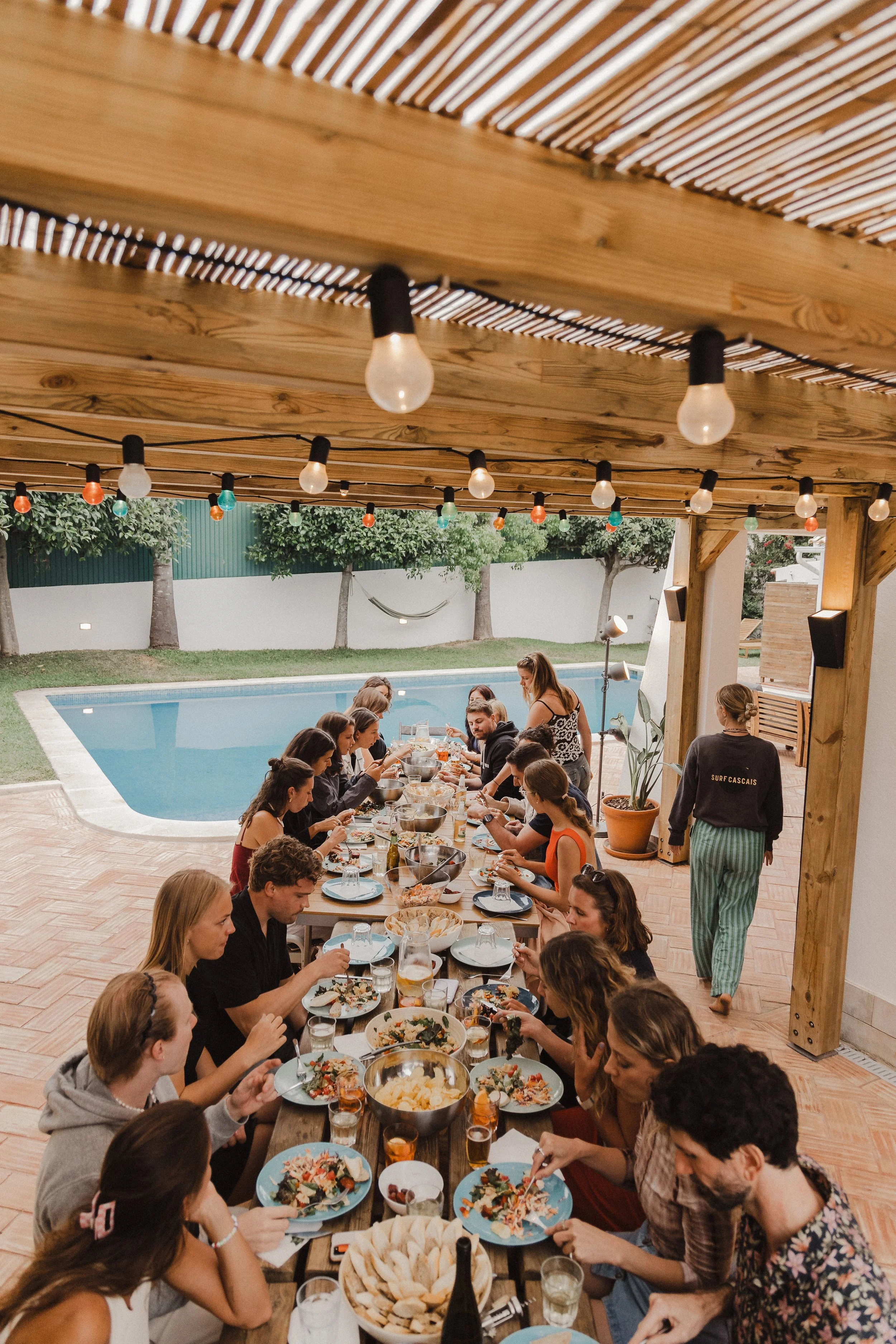 People gathered around a long table enjoying a meal outdoors near a swimming pool, under a wooden pergola with string lights.