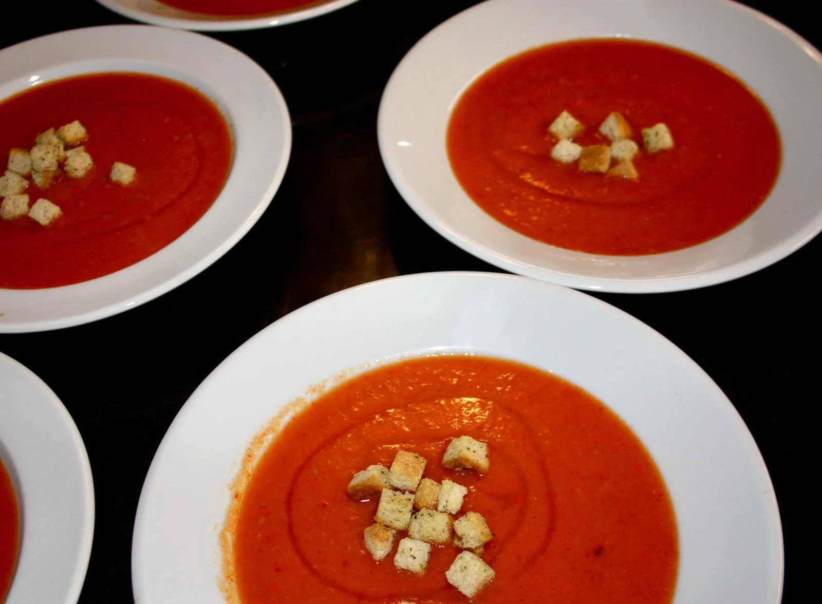 Three white bowls filled with tomato soup, topped with croutons, on a black surface.