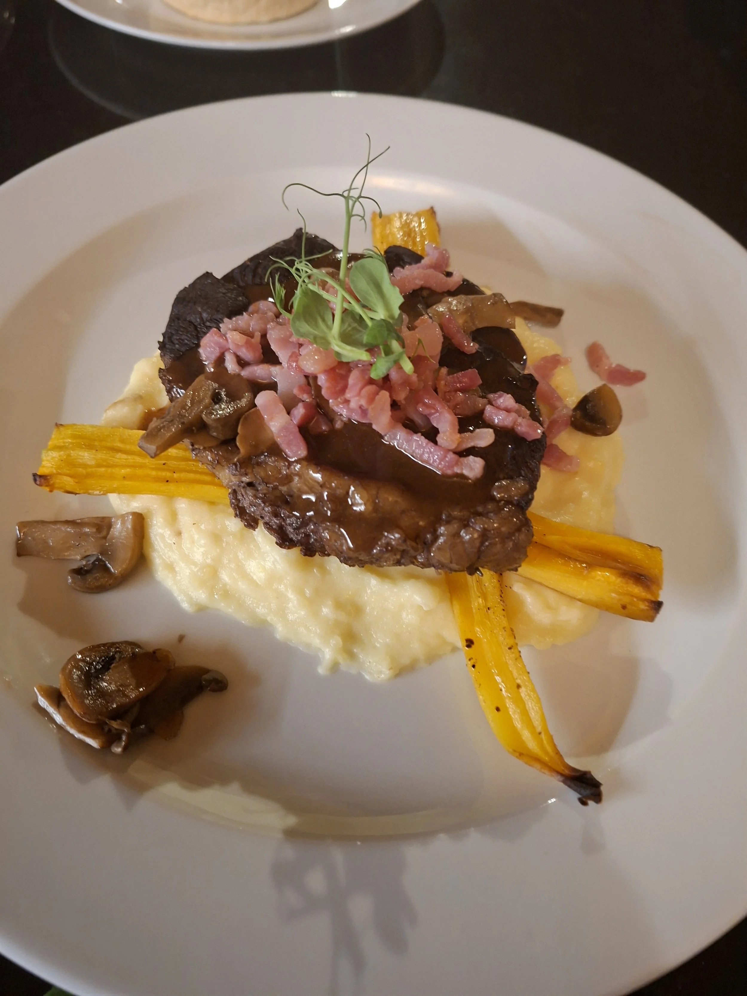 A plated dish featuring mashed potatoes, grilled vegetables, topped with crispy bacon bits, a sprig of greenery, and a charred yellow pepper, with a side of cooked mushrooms.