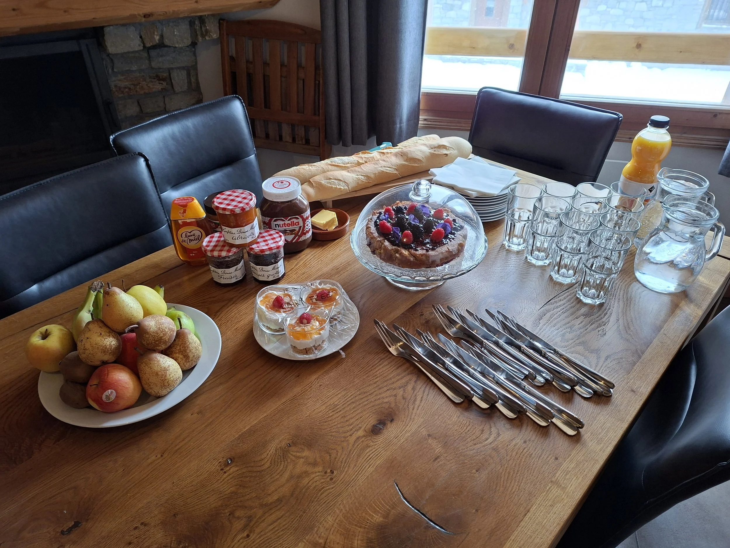 A wooden table set for breakfast with apples, a large baguette, jars of jam, a cake with berries, a bottle of orange juice, multiple empty glasses, and cutlery. There are plates with small desserts and a glass pitcher of water. The background shows a window, curtains, and part of a stone fireplace.