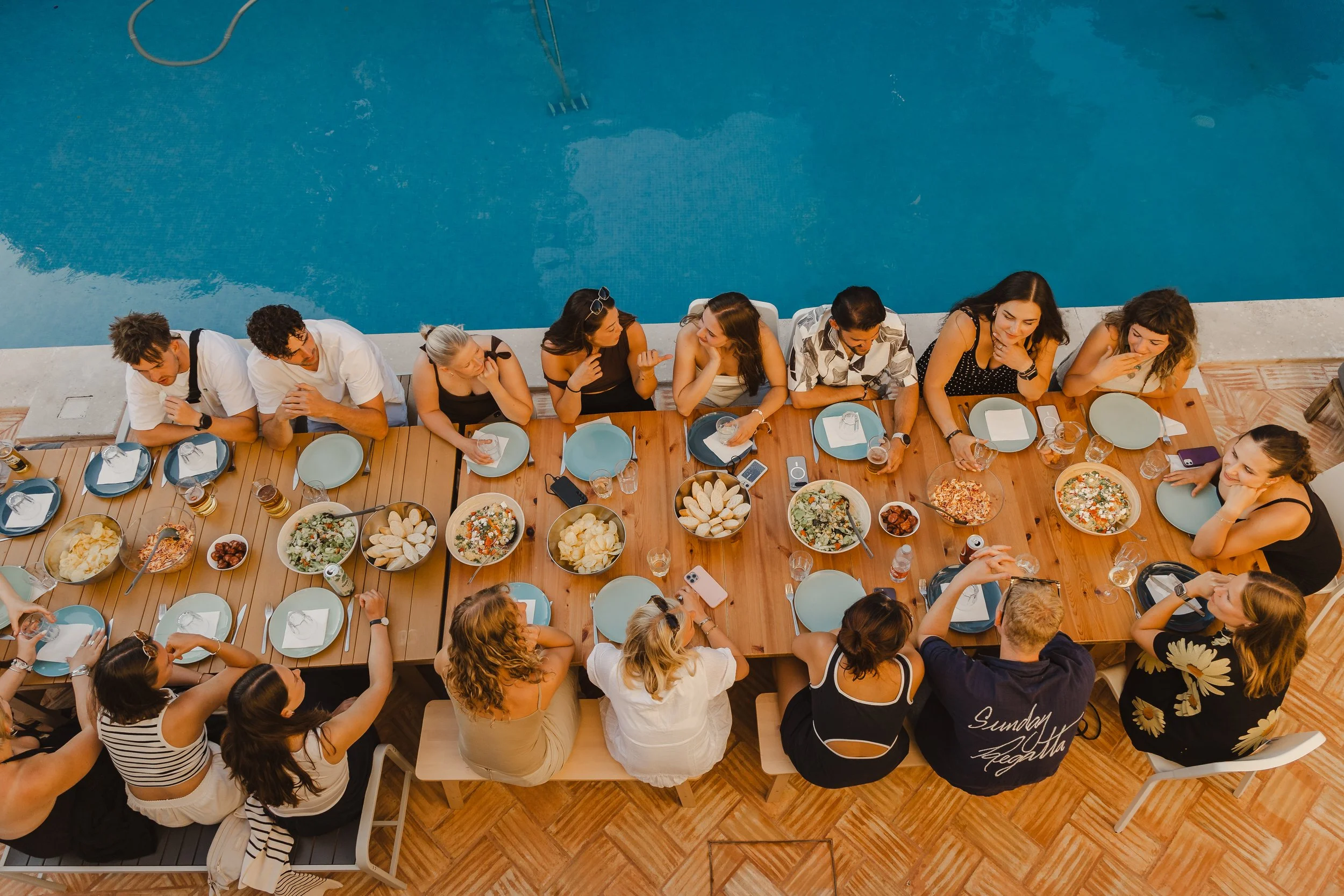 A group of people sitting at a long dining table outdoors next to a swimming pool, enjoying a meal with pizza, salads, and drinks.