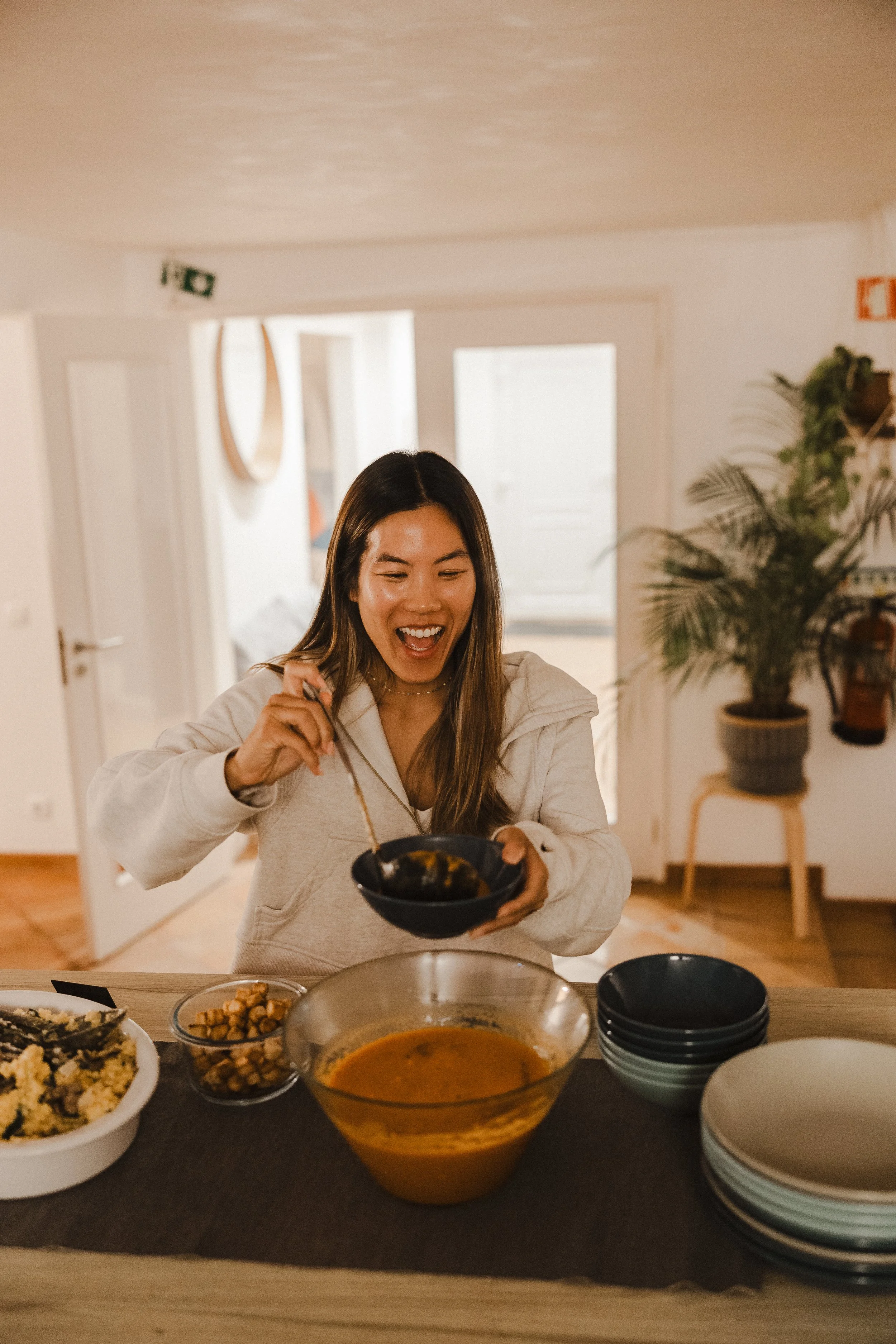 A woman with long brown hair wearing a white hoodie is smiling and holding a bowl of food in her left hand and a spoon in her right hand. There are various bowls and dishes of food on the table in front of her, including a bowl of soup, a bowl of chi