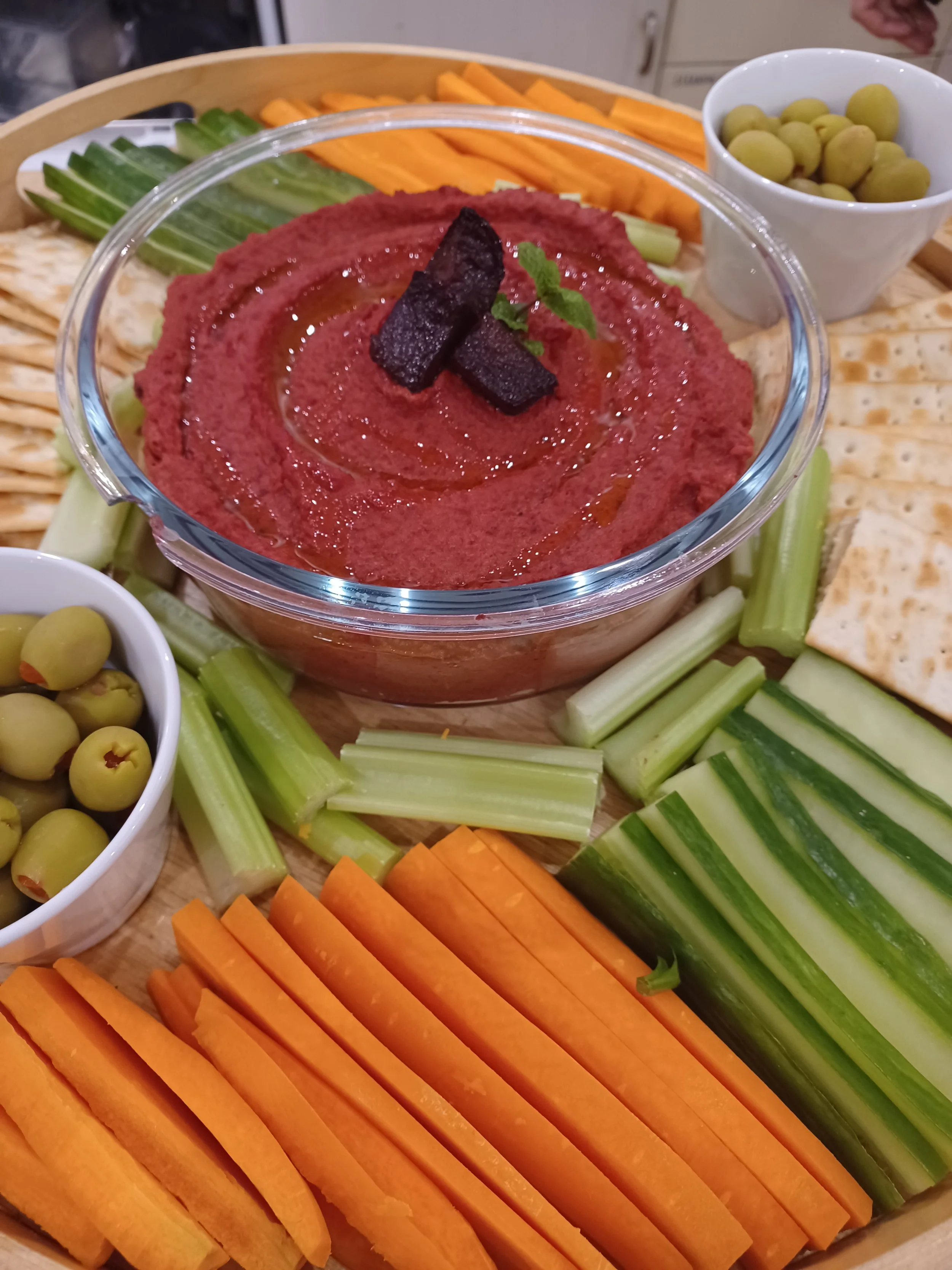A platter containing various fresh vegetables and dips, including sliced carrots, celery sticks, green olives, and a bowl of red hummus with a cherry tomato and a small green herb garnish on top.