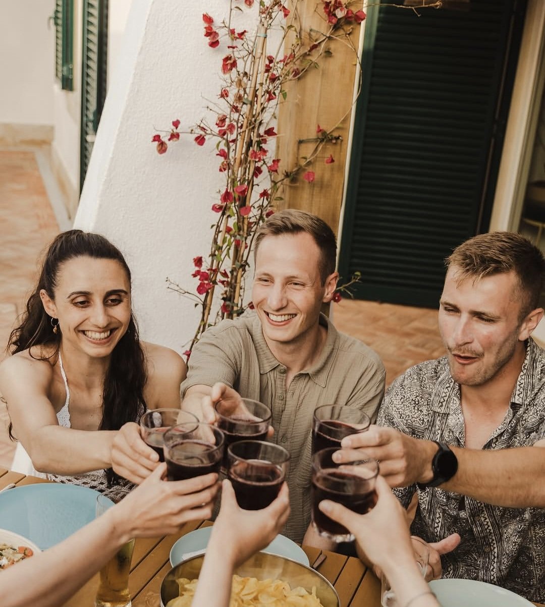 People toast with glasses of red wine at a dinner party outdoors, smiling and enjoying each other's company.