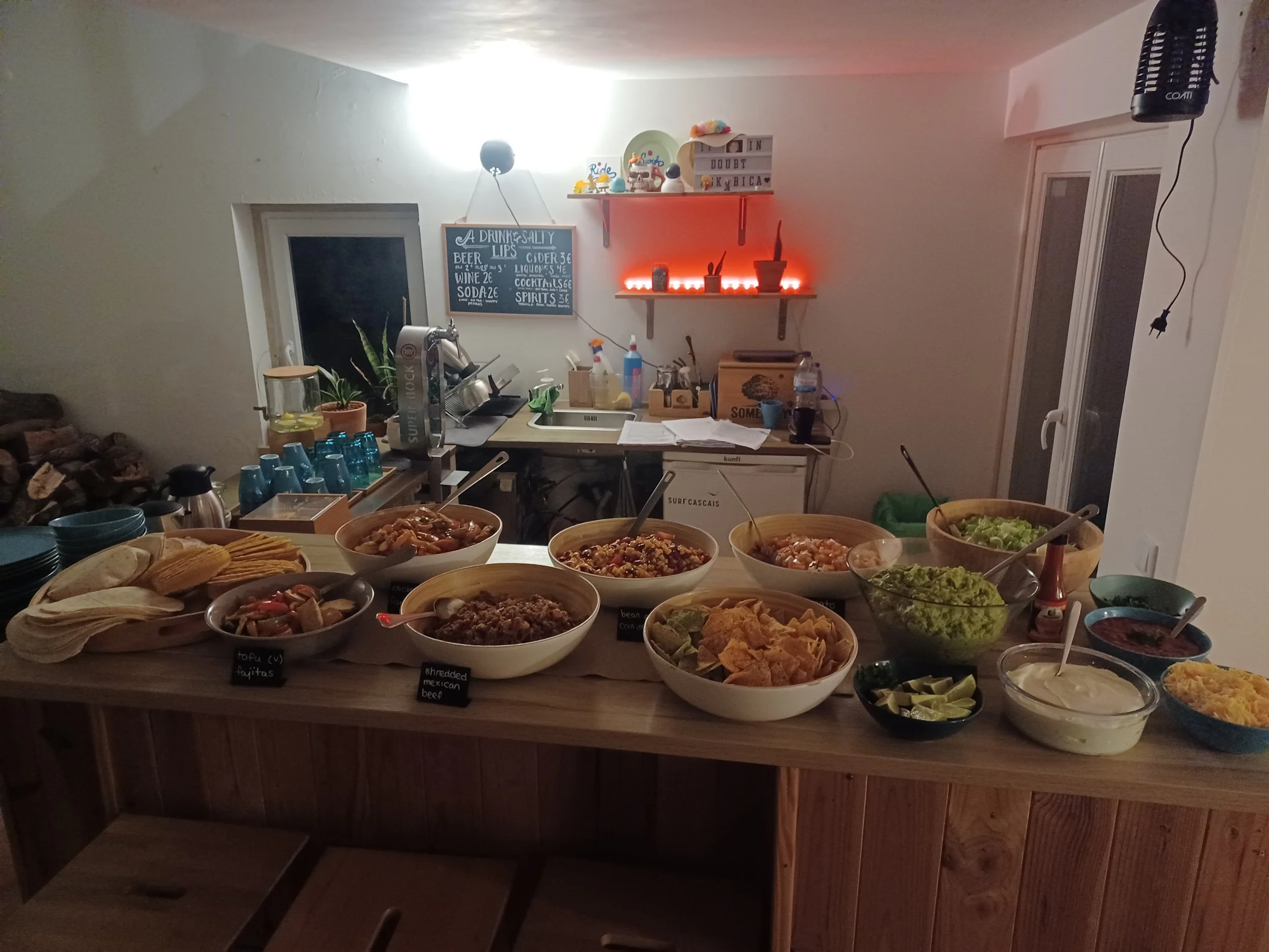 A kitchen counter filled with various bowls of food including chips, salsa, guacamole, and other dips, with a background of kitchen shelves and a chalkboard menu.
