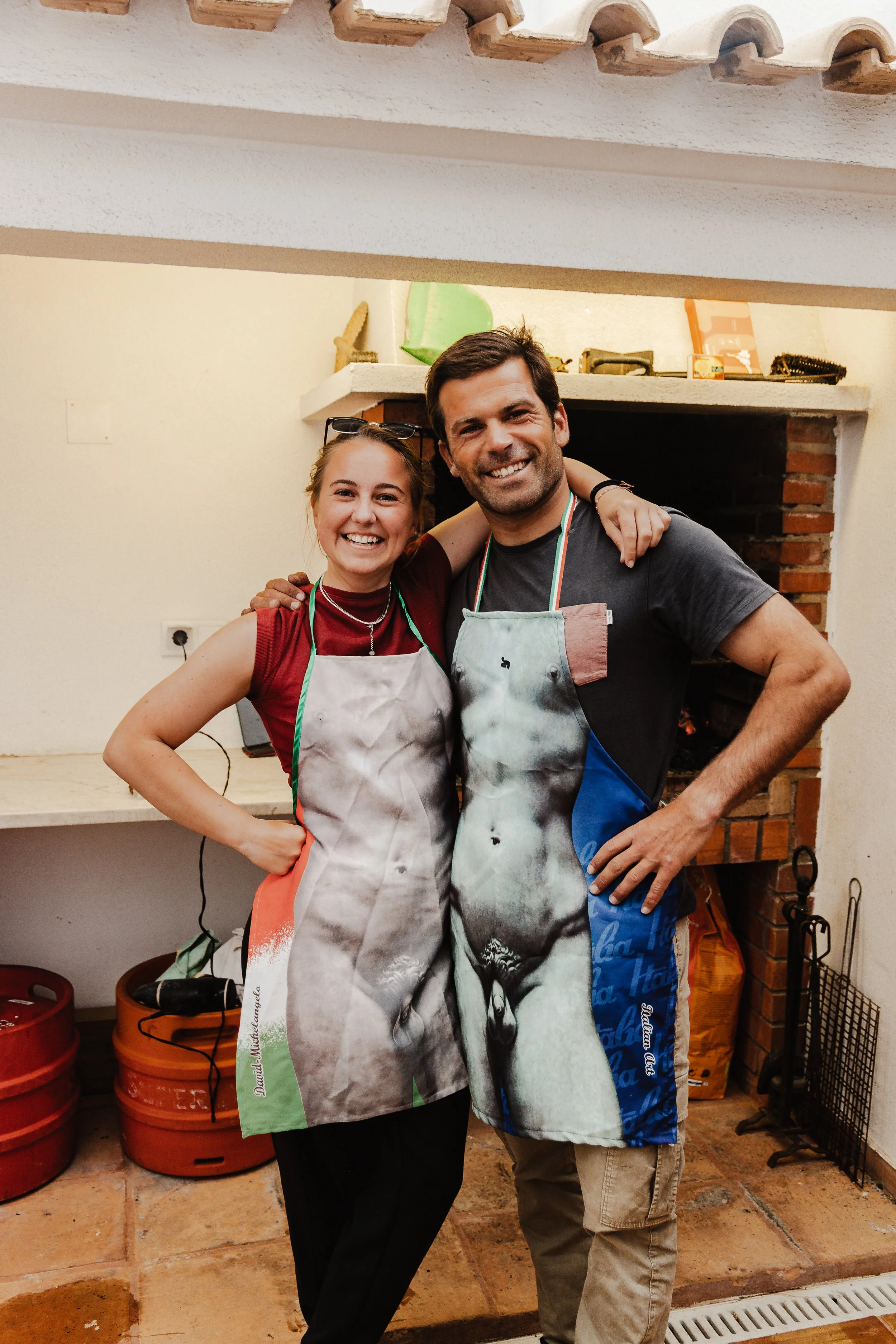 Two smiling people, a woman and a man, standing together indoors in front of a fireplace, wearing aprons with animal prints, with their arms around each other.