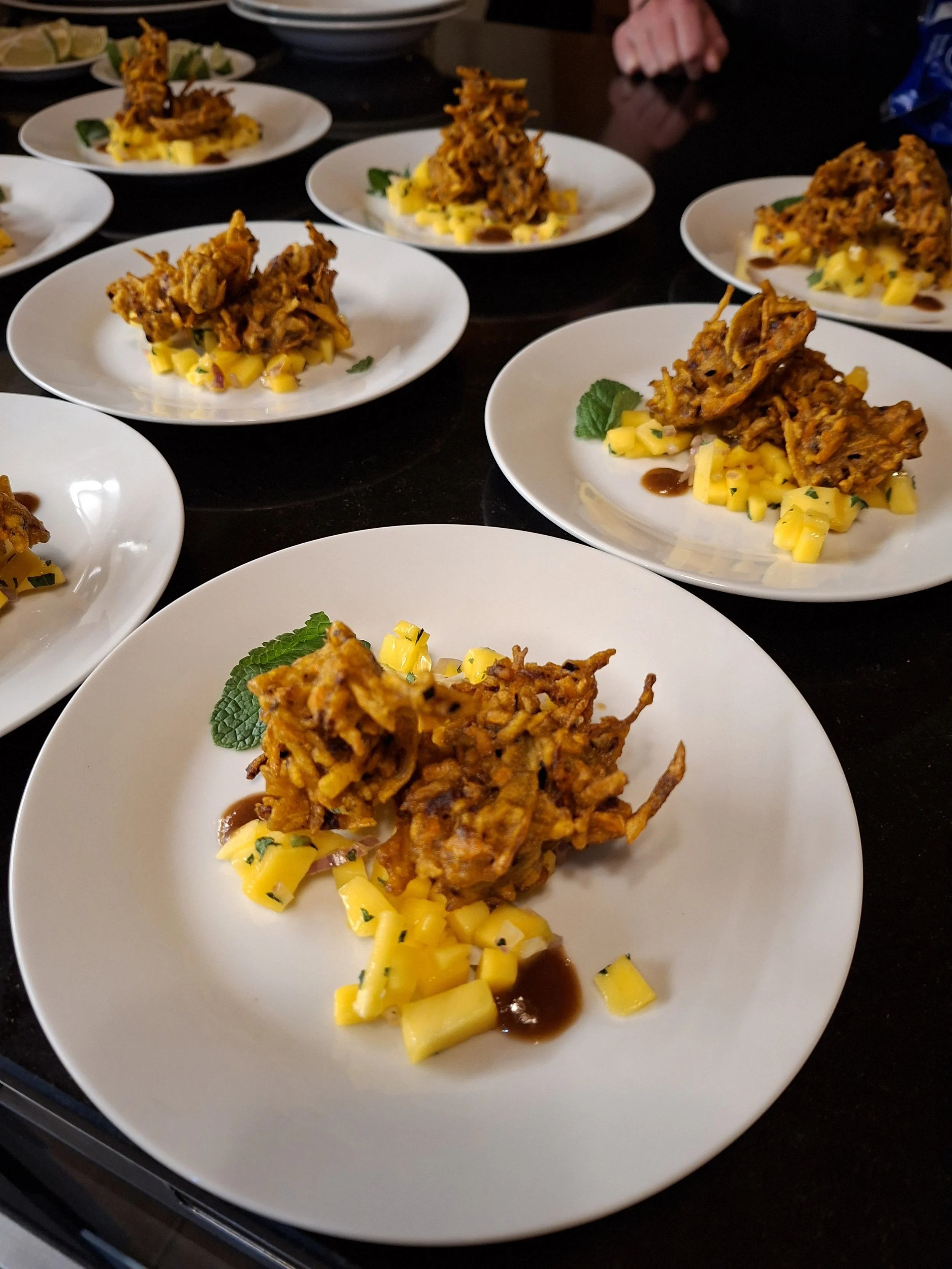 Plates of Indian dish, likely pakoras or fritters, served with mango chunks, green leaves, and a brown sauce.