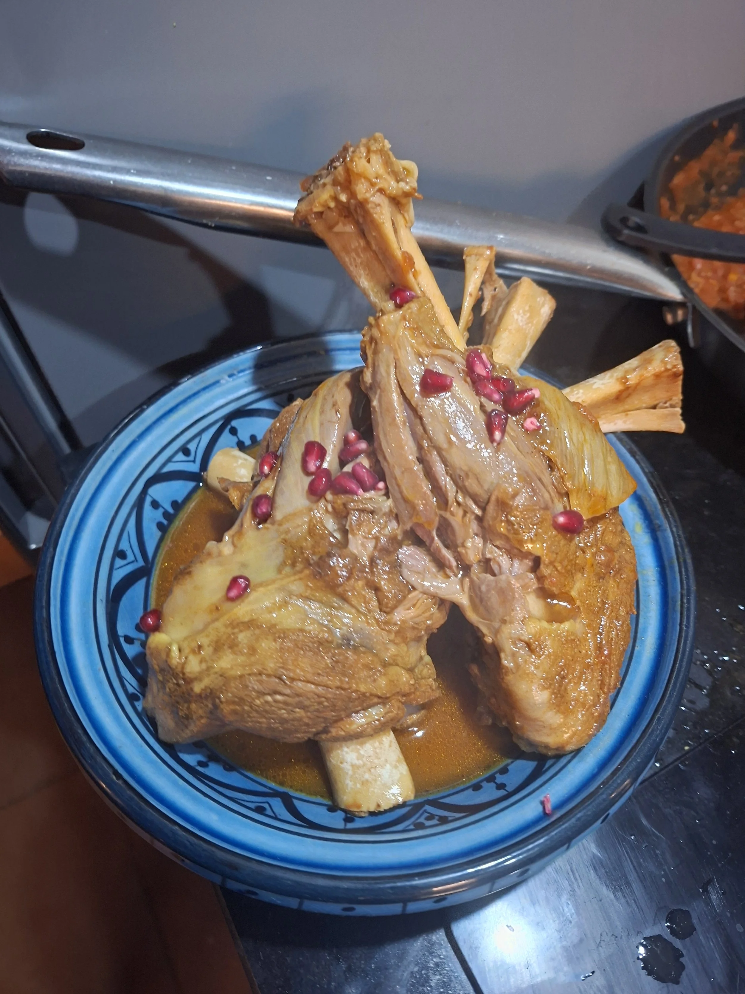 Cooked meat with bones in a blue patterned bowl, topped with pomegranate seeds, on a dark surface with some water droplets.