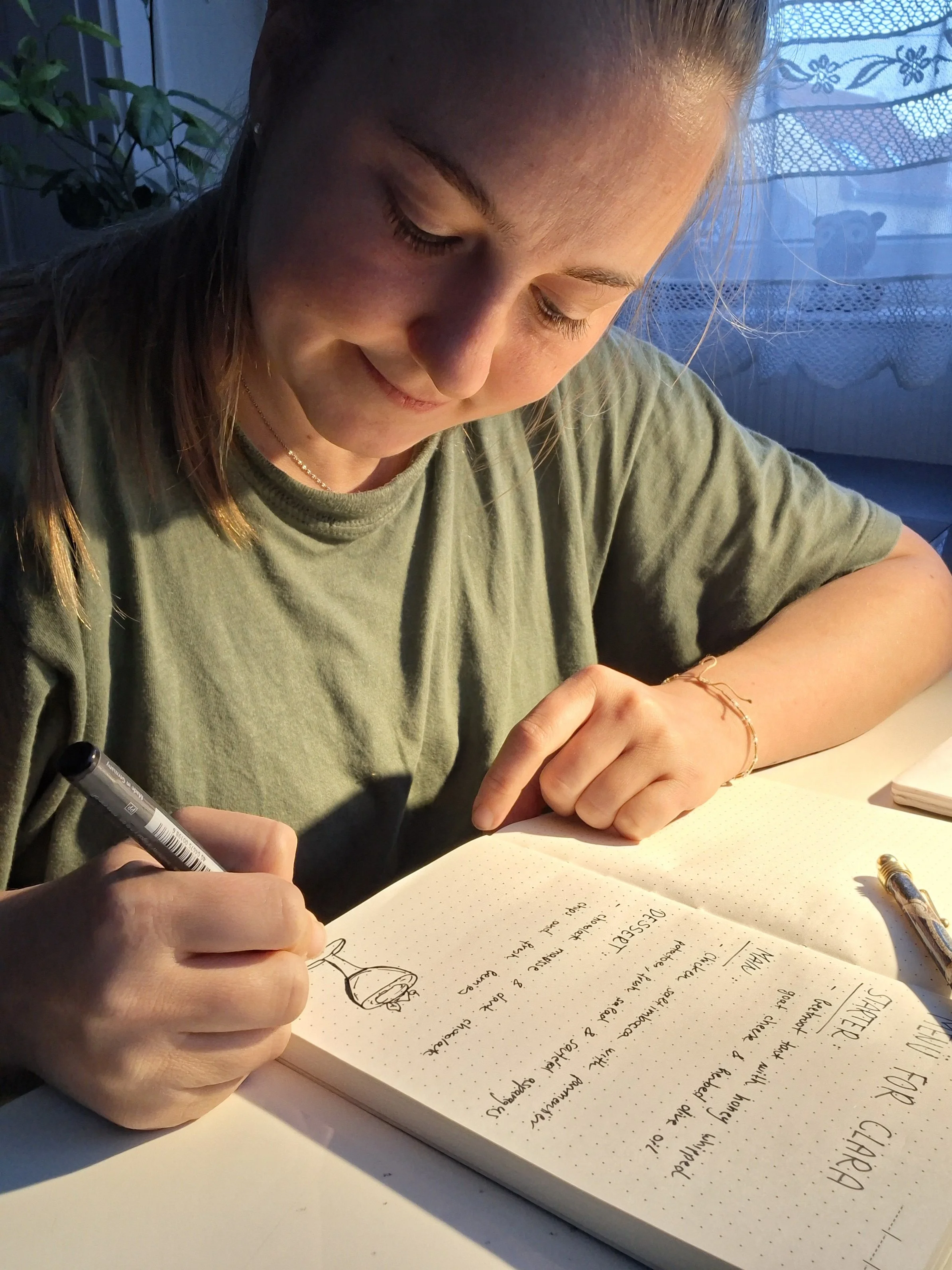 A person with long hair, wearing a green T-shirt, is sitting at a table and writing in a notebook with a black pen. The notebook contains handwritten notes and a drawing. There is a silver pen on the table, and in the background, a window with lace curtains and a potted plant are visible.