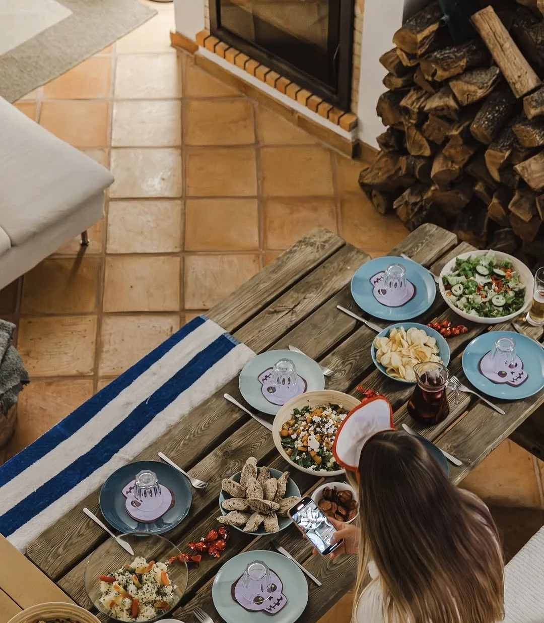 A wooden dining table set with various dishes, including salad, bread, and desserts, in a cozy living room with tiled floors, a stacked woodpile, and a woman taking a photo of the food.