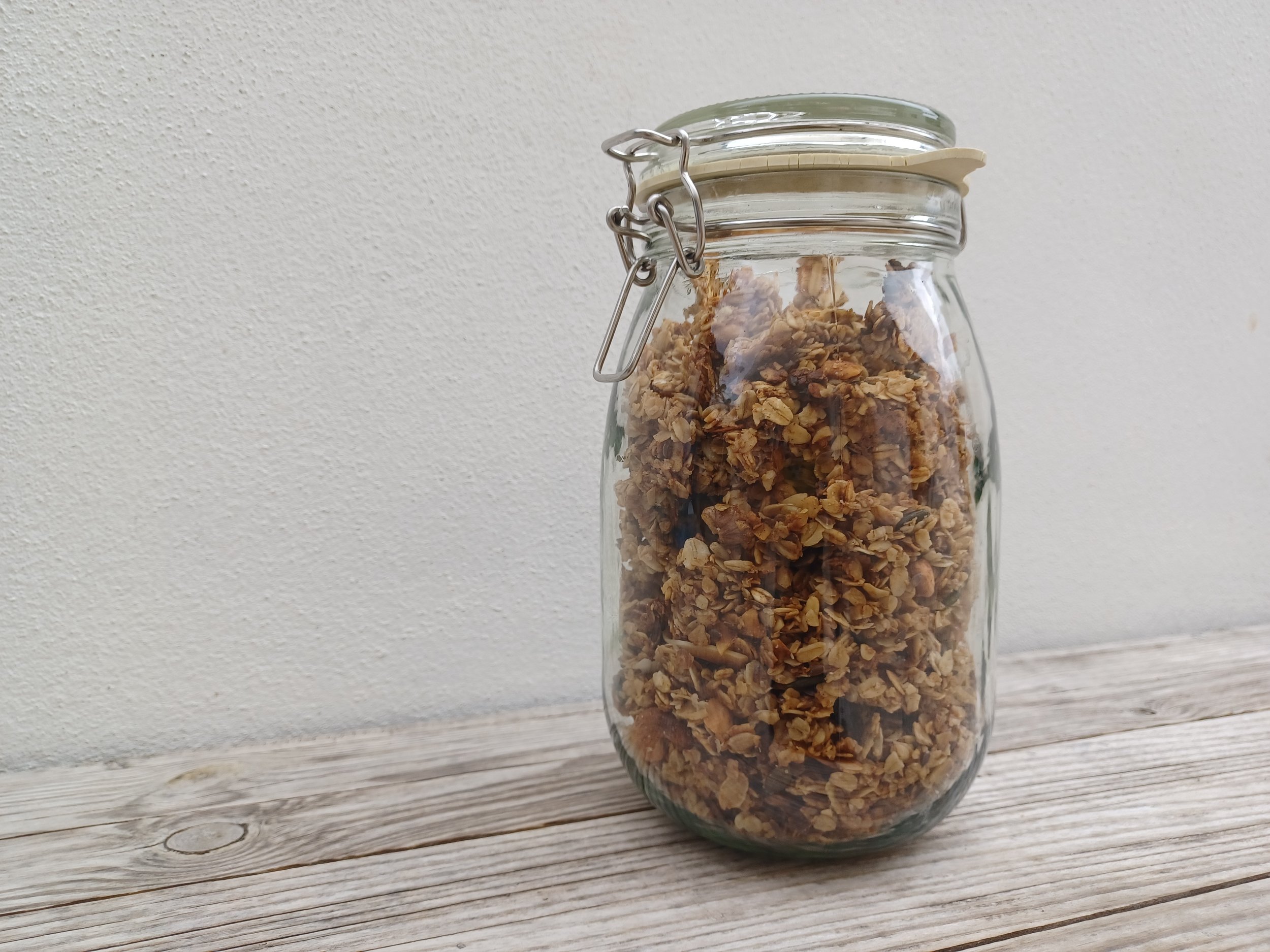 A glass jar filled with granola sits on a rustic wooden surface, with a plain white wall in the background.