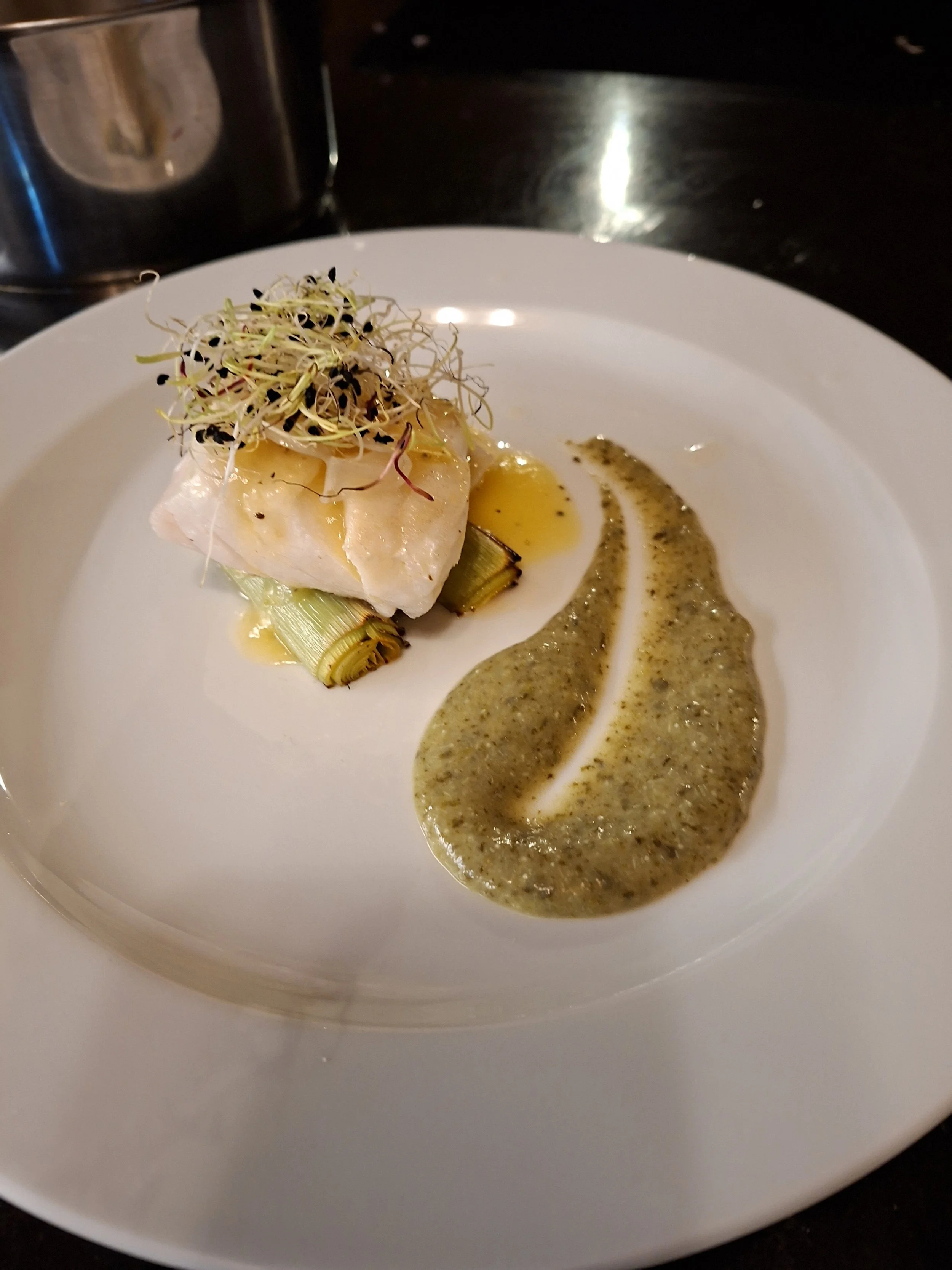 A plated dish with a piece of fish topped with microgreens, served with a sauce on the side.