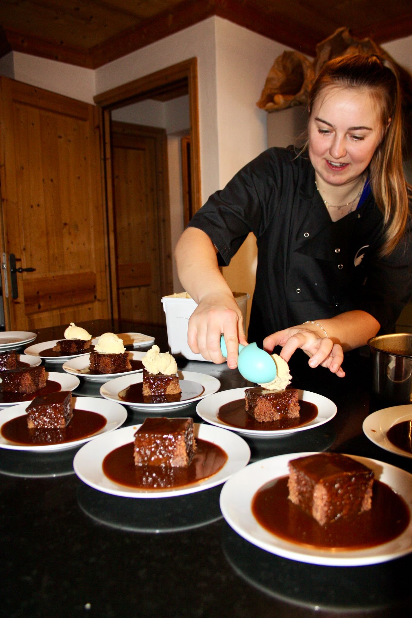 A woman in a black shirt decorates a chocolate cake with a dyed blue egg-shaped decoration while surrounded by plates of chocolate cake with caramel and whipped cream topping on a dark table.