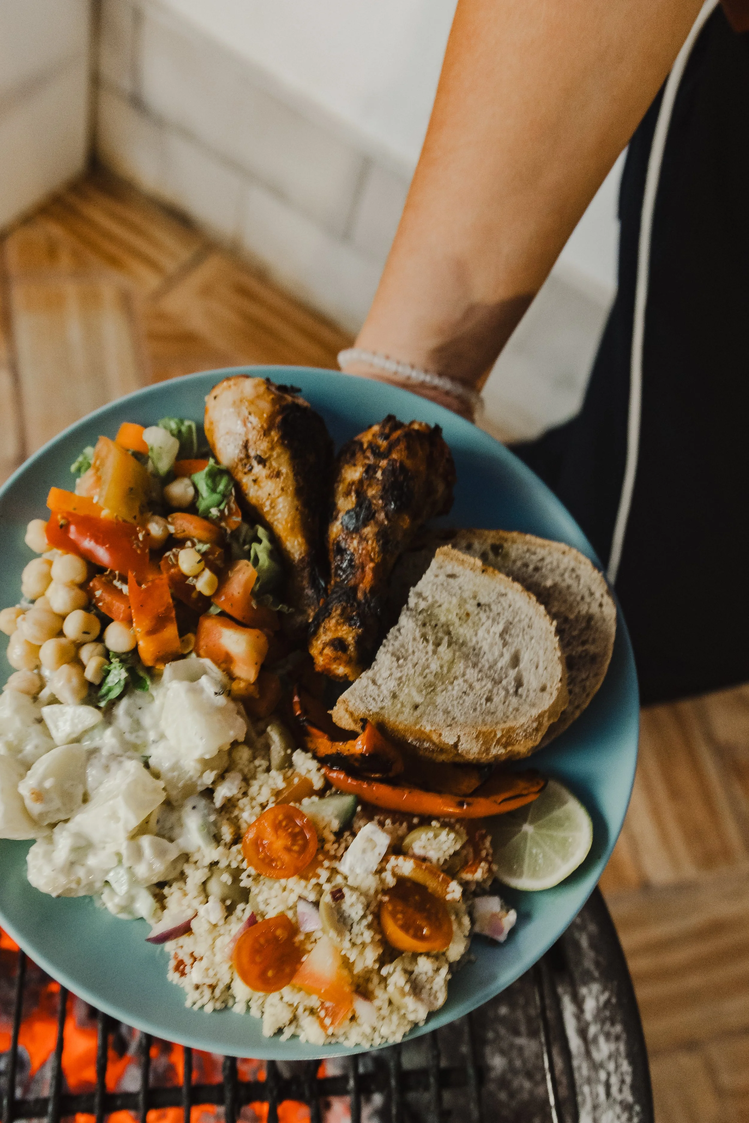 A plate of food with grilled chicken, salad, corn, bread, and lemon wedges, set on a grill over an open flame.