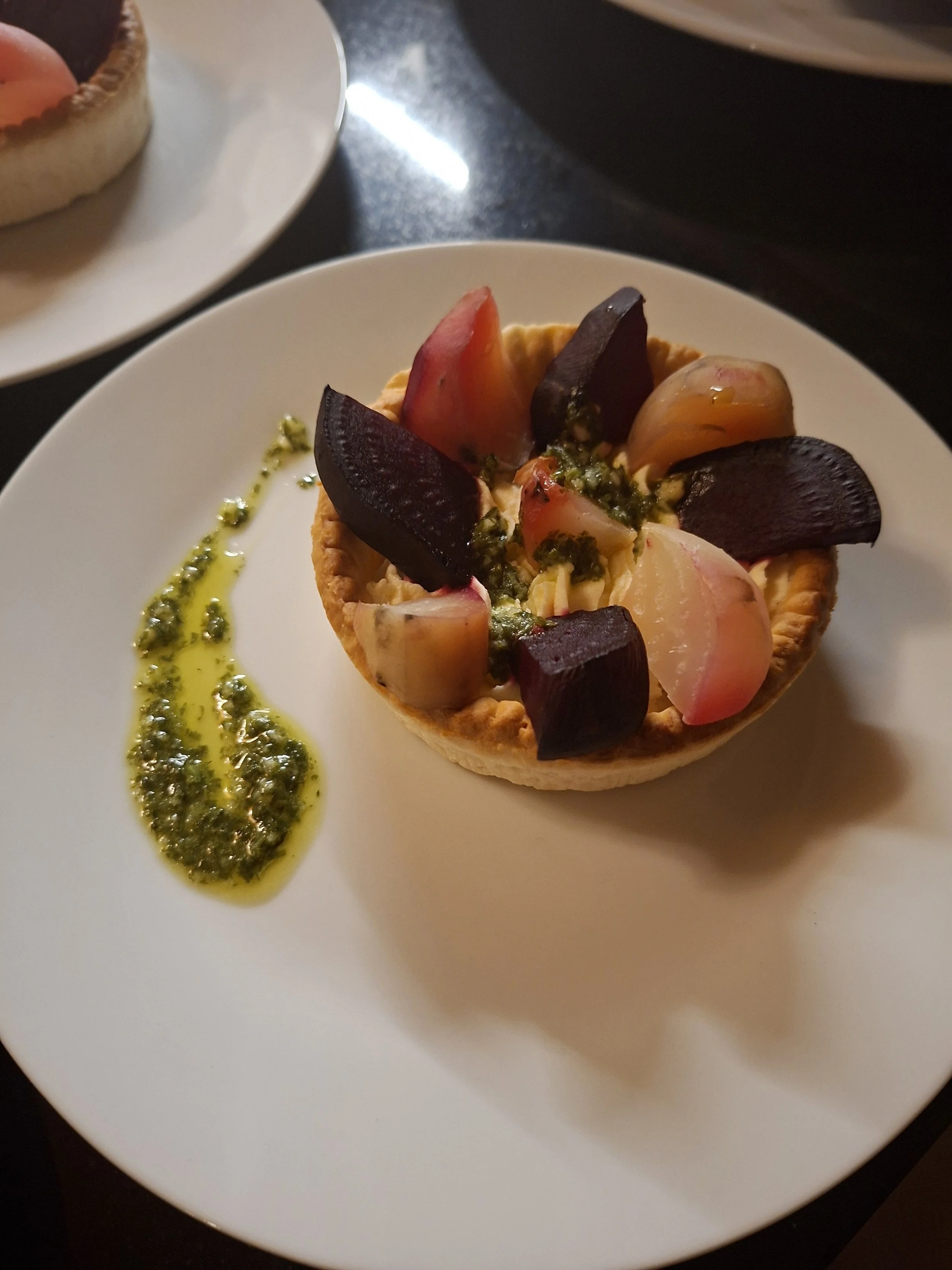 A tart with a golden crust filled with various fruits, topped with green sauce, served on a white plate with a drizzle of green sauce alongside.