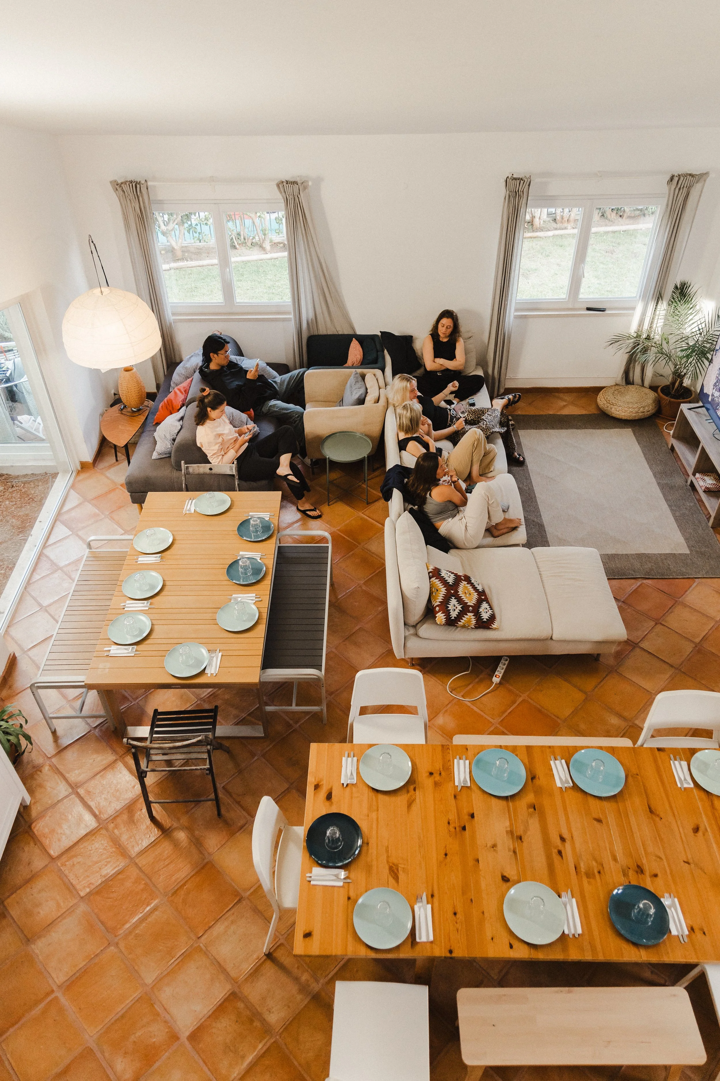 A living room with tiled floors, a large wooden dining table set with light blue plates and cutlery, a beige sofa with people seated, a black sofa, and large windows with curtains, overlooking a garden with grass and trees.