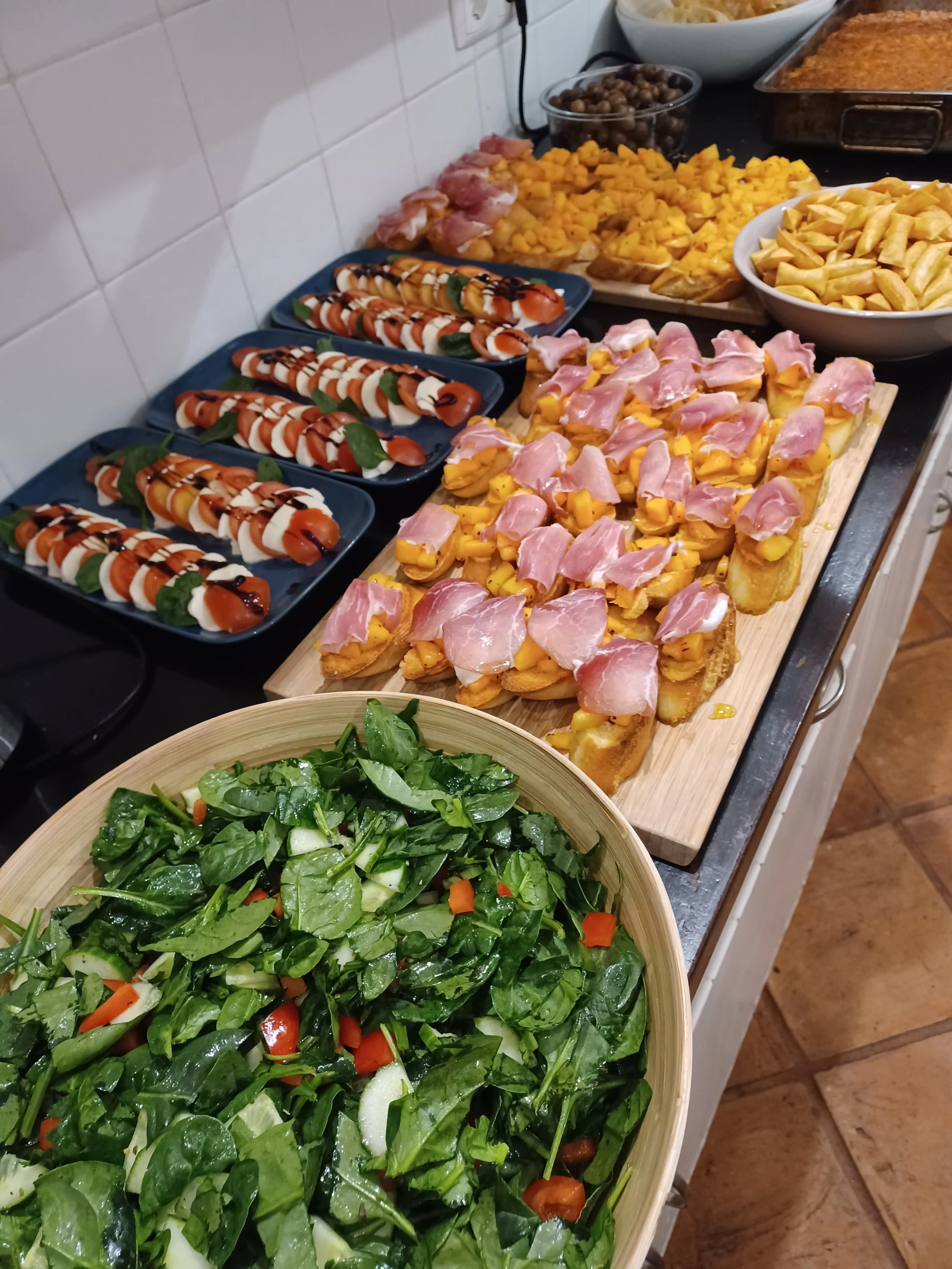 A kitchen counter with various prepared foods, including a large fresh green salad, small Caprese salads with basil, a tray of fried chicken topped with ham, bowls of potato chips, chunks of cooked sweet potatoes, and a tray of baked casserole.