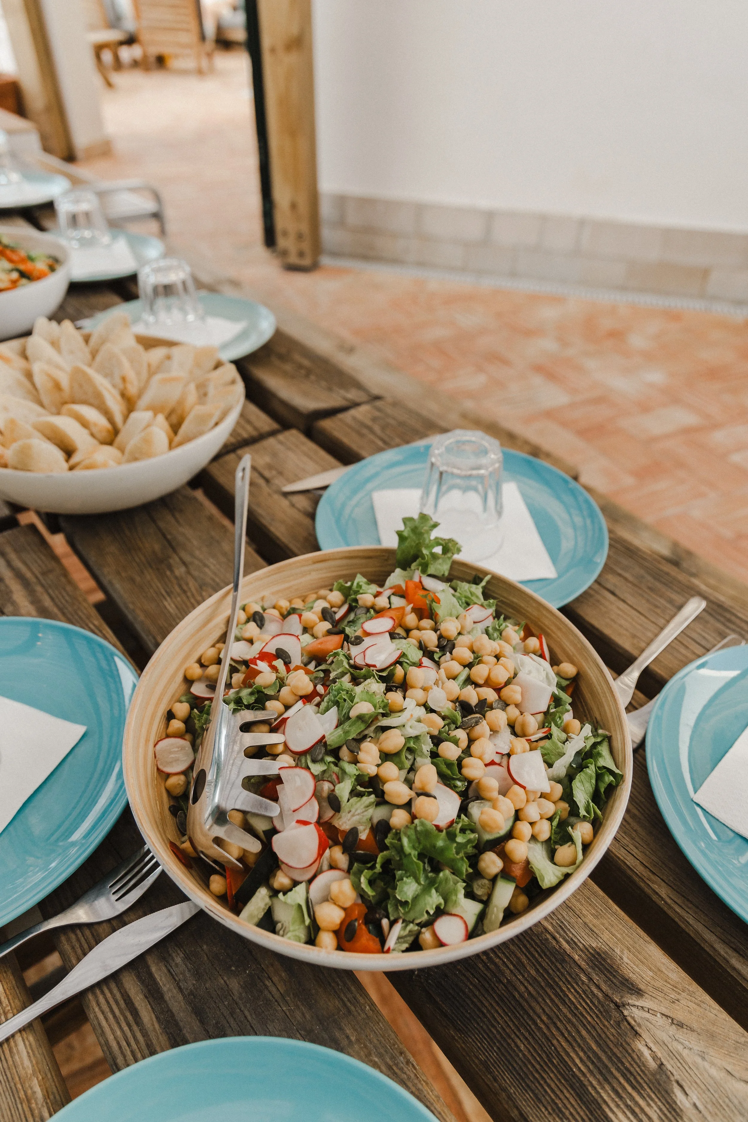A wooden table with bowls and plates of salad and chips, set for a meal, with upside-down glasses and utensils on paper napkins in a rustic dining setting.