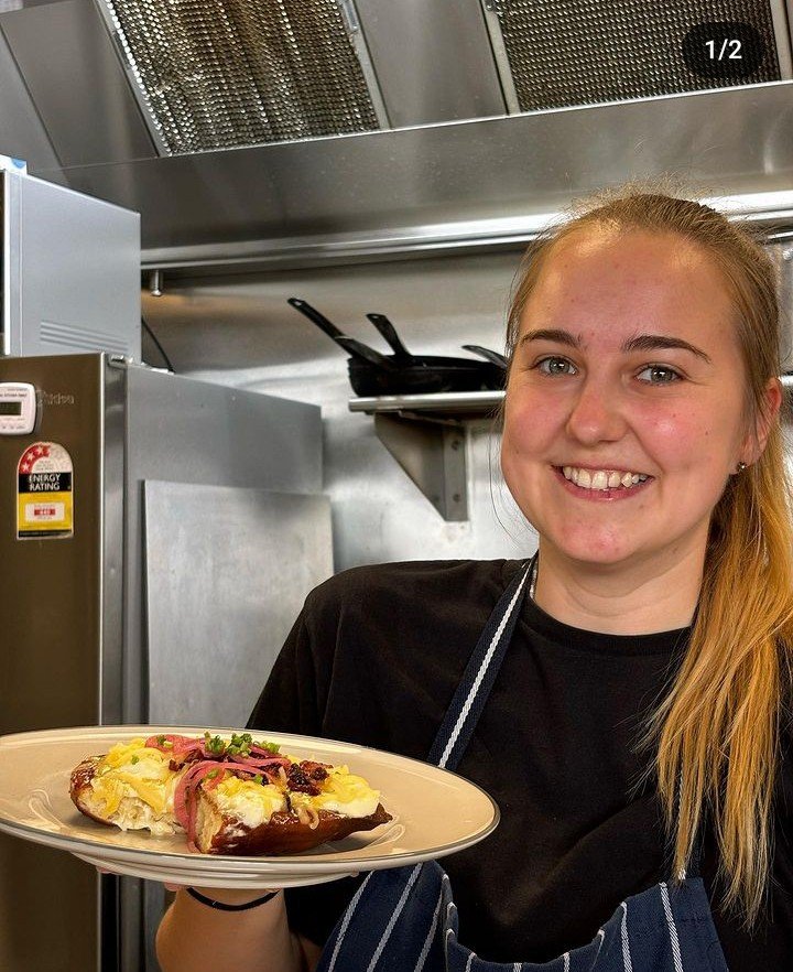 A young woman with blonde hair and a black shirt holding a plate with a slice of loaded pizza, smiling in a commercial kitchen.