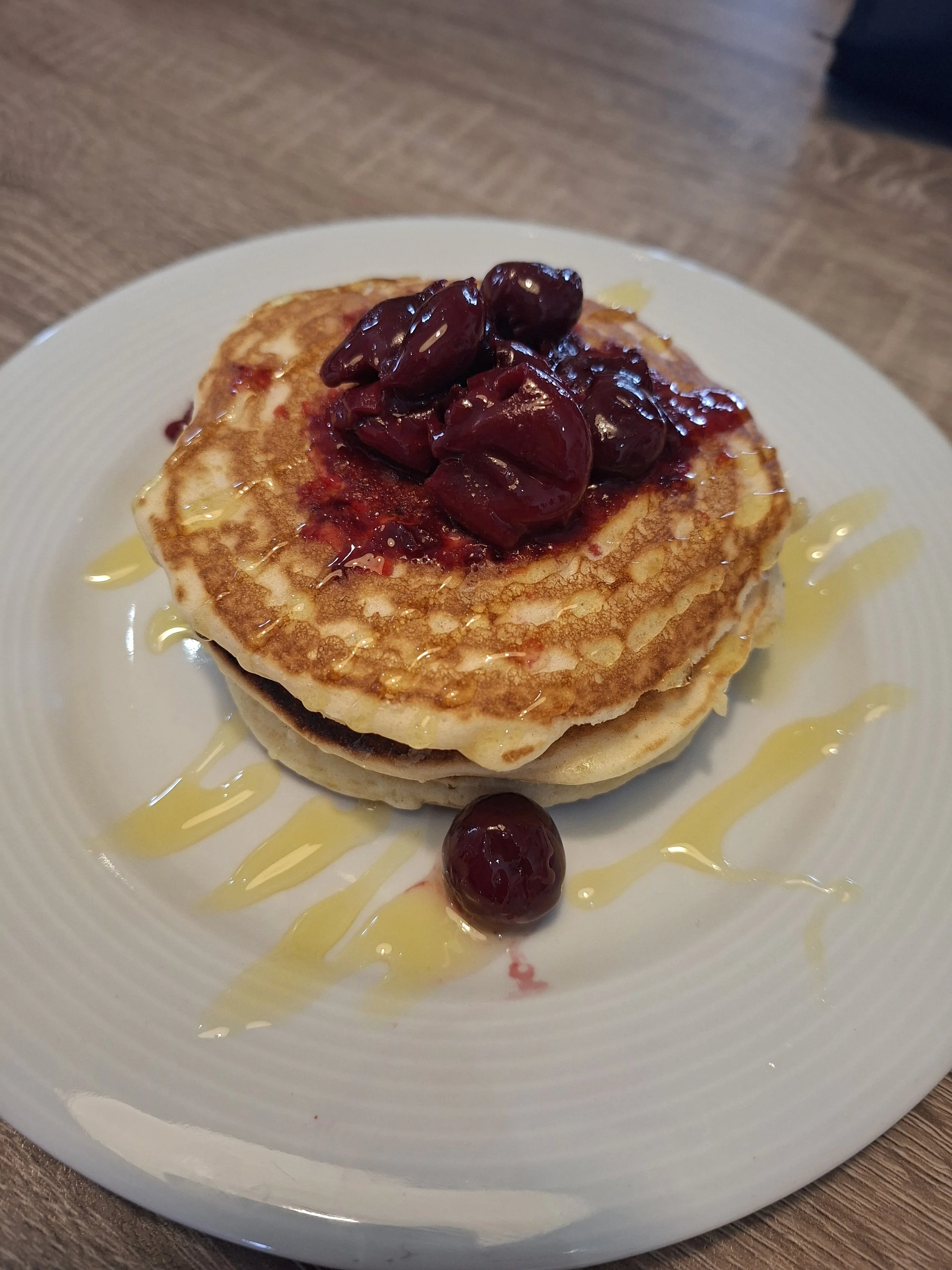 Stacked pancakes topped with dark cherries, drizzled with syrup and honey, served on a white plate with a cherry and sauce drizzle.