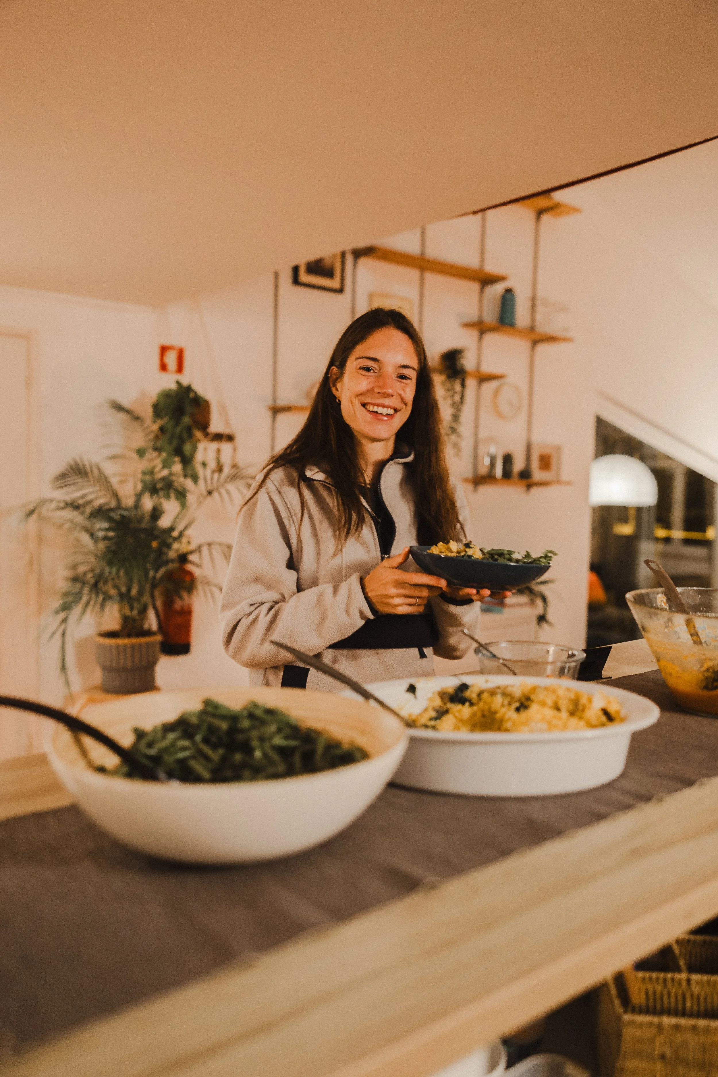 A young woman with long brown hair smiling while holding a bowl of food in a cozy, well-lit kitchen or dining area with decorative shelves and houseplants.