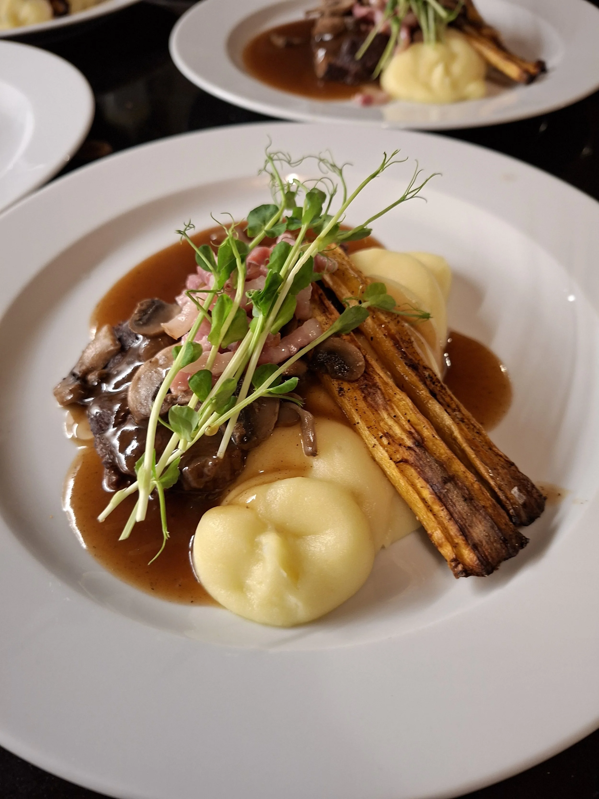 A plated dish featuring a crispy fried vegetable, topped with microgreens, alongside creamy mashed potatoes, with mushroom gravy and sautéed mushrooms.