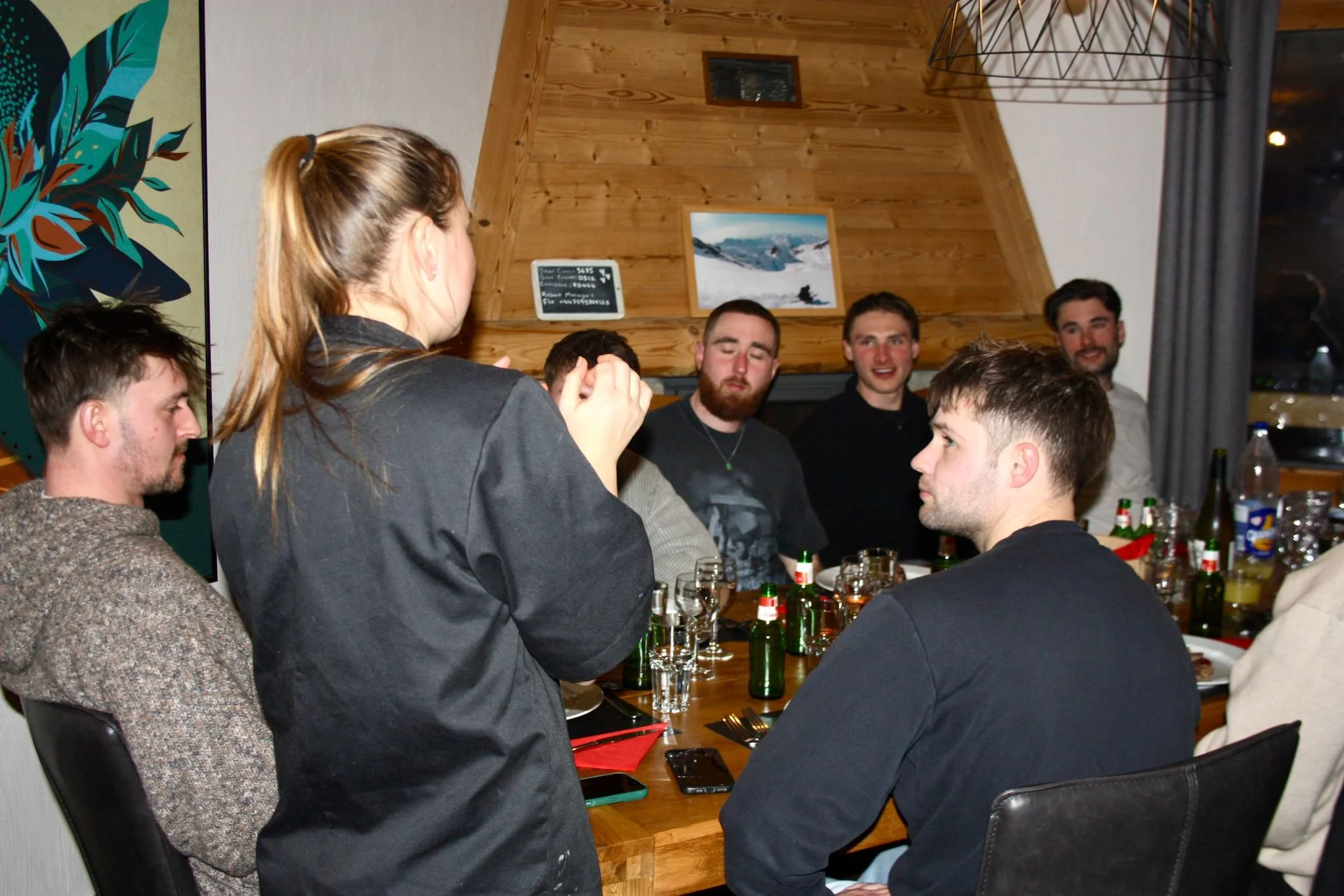 A group of young adults gathered around a wooden table, with some standing and others seated, in a cozy indoor setting with wood-paneled walls. They are socializing, with drinks and snacks on the table, and a woman standing appears to be speaking or 