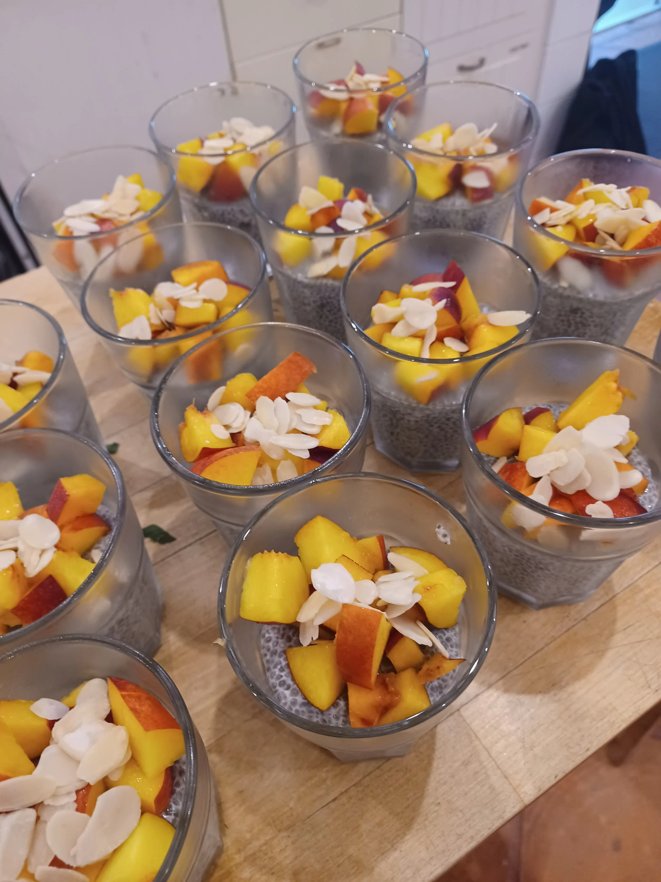 Multiple glasses filled with chia pudding topped with sliced peaches and almond flakes, arranged on a wooden surface.