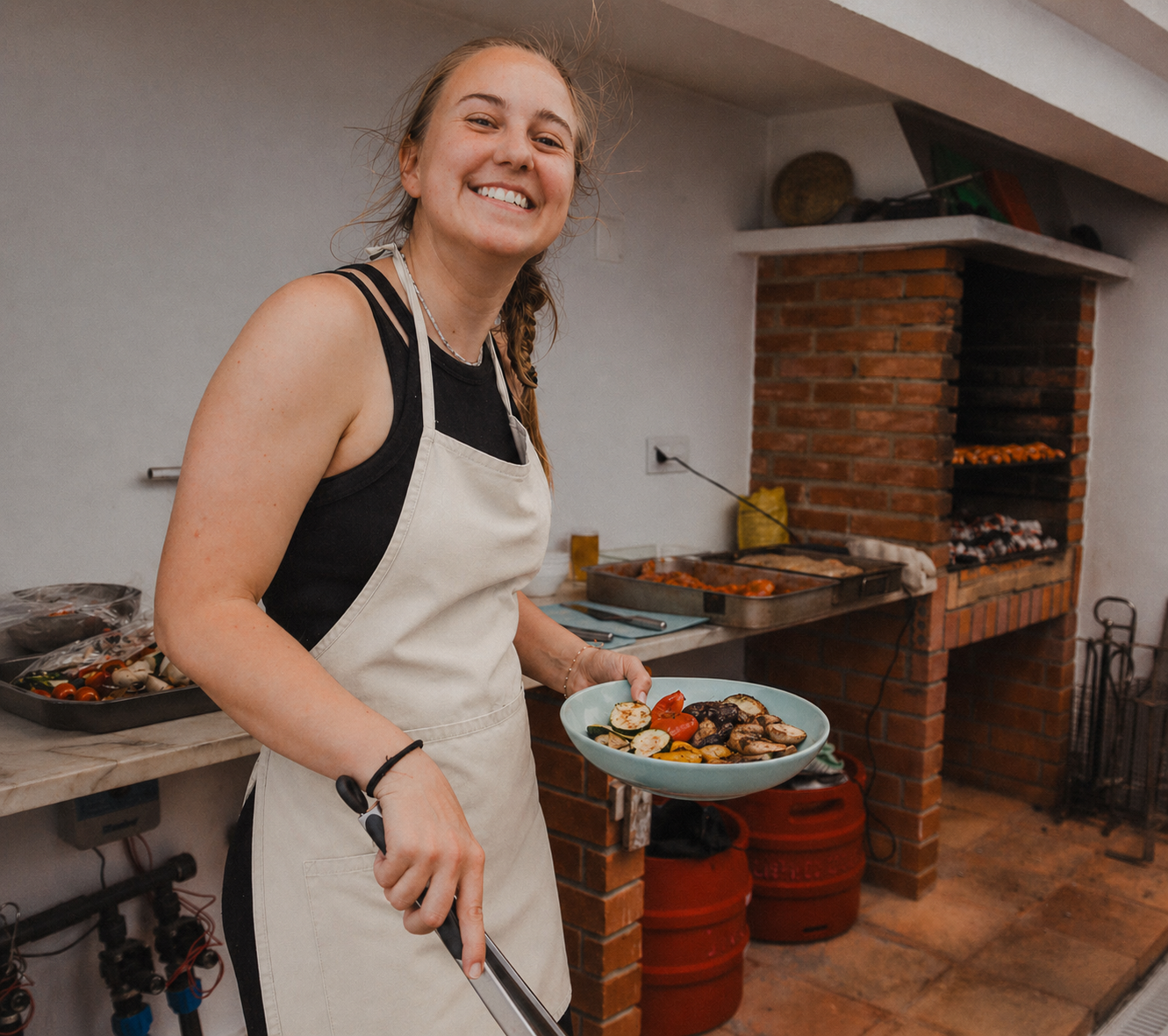 A woman smiling while holding a plate of cooked vegetables, standing in a kitchen near a brick grill with food grilling inside.