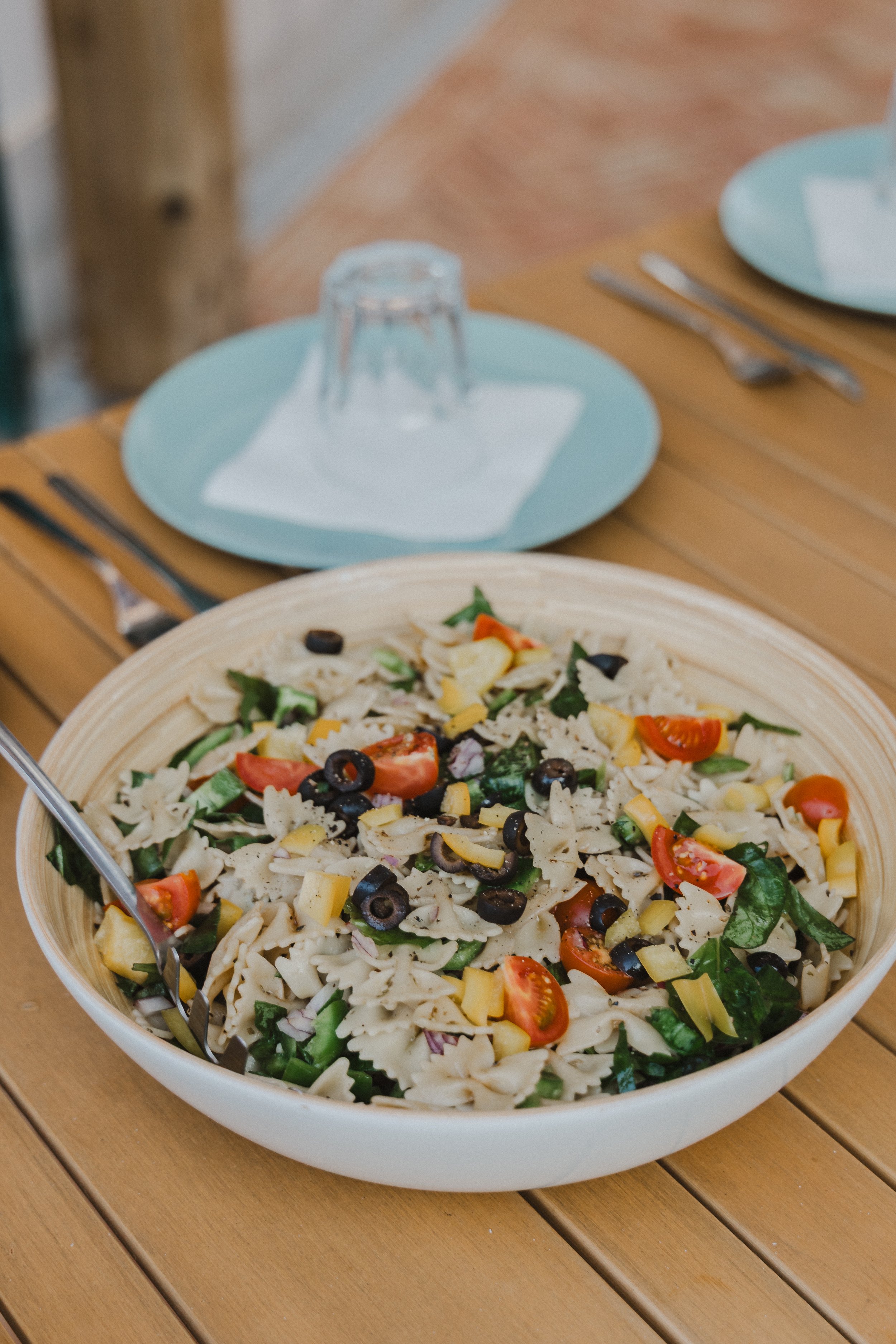 A large bowl of pasta salad containing cherry tomatoes, black olives, chopped spinach, and cheese, placed on a wooden table.