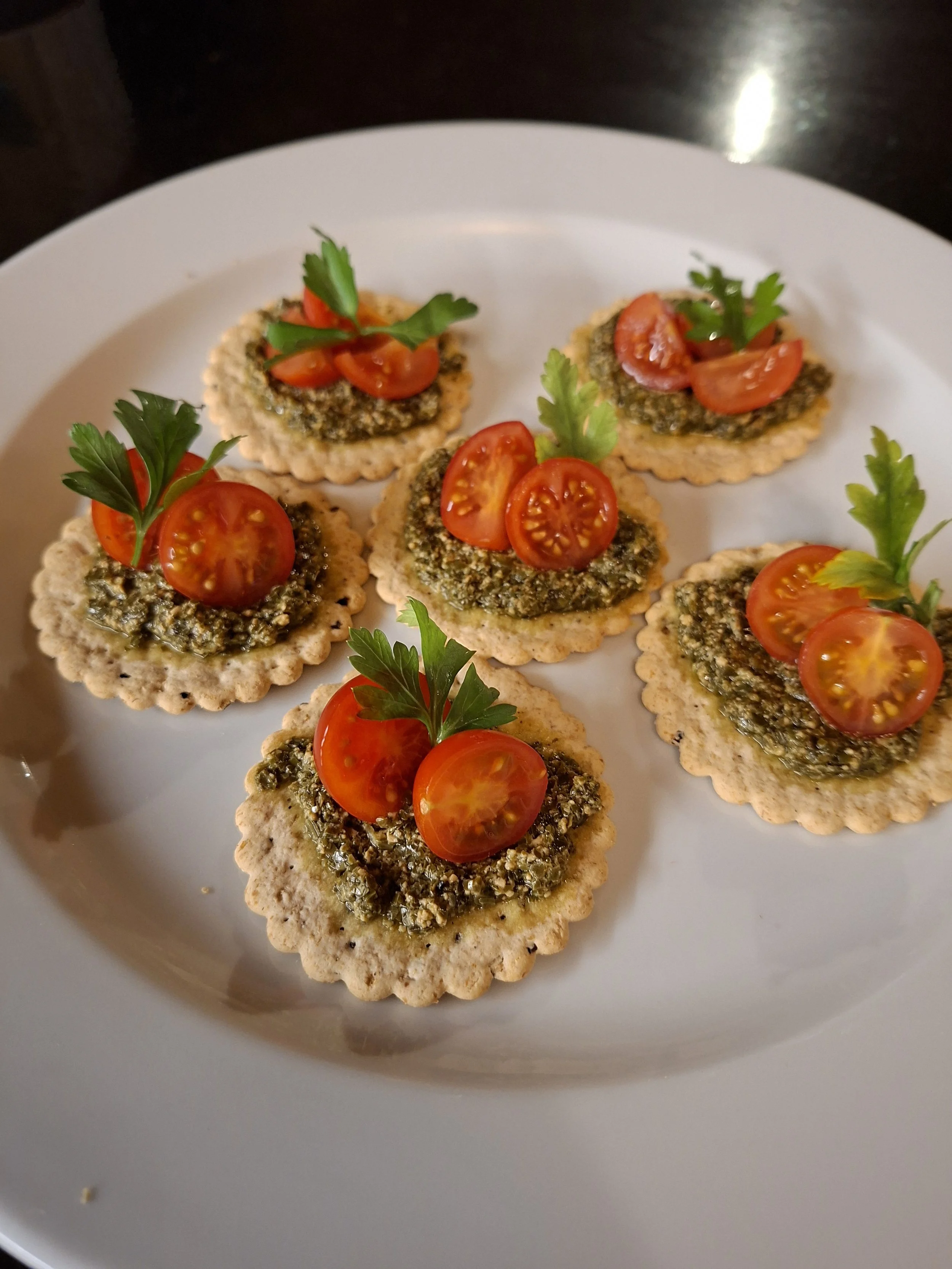 Crackers topped with a green pesto, halved cherry tomatoes, and fresh parsley on a white plate.