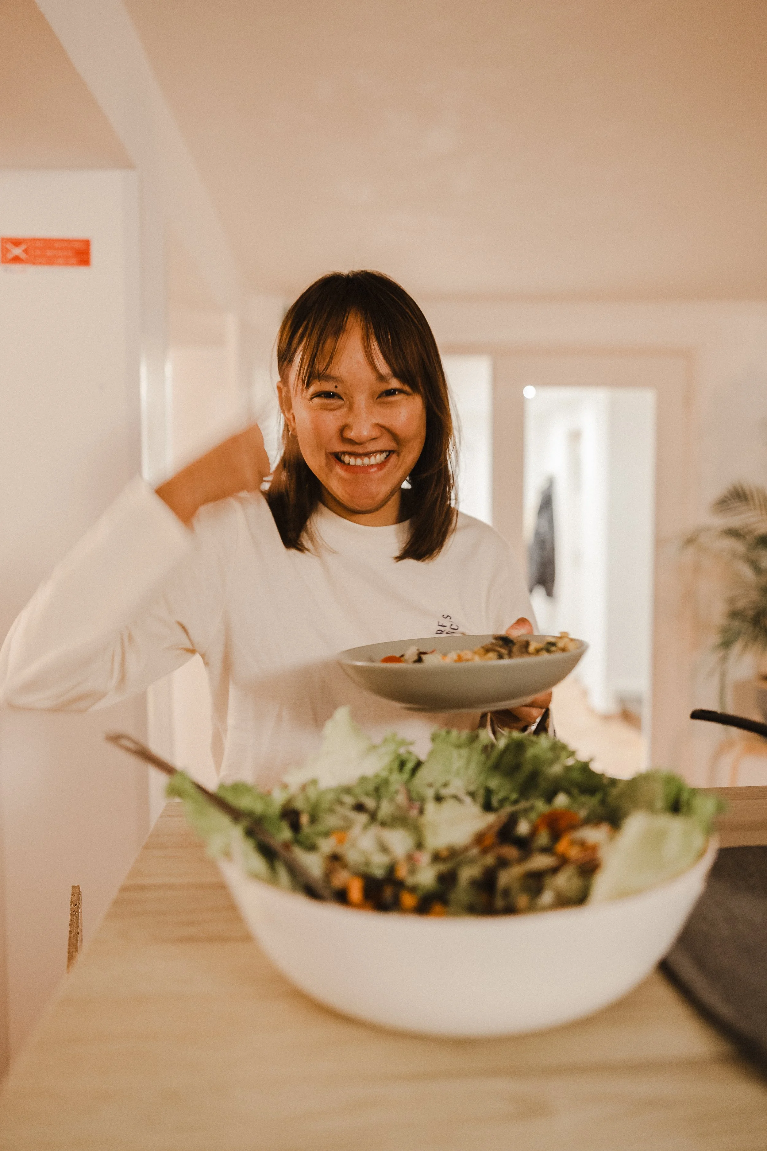 A woman with shoulder-length hair smiling and holding a plate of food in a brightly lit room.
