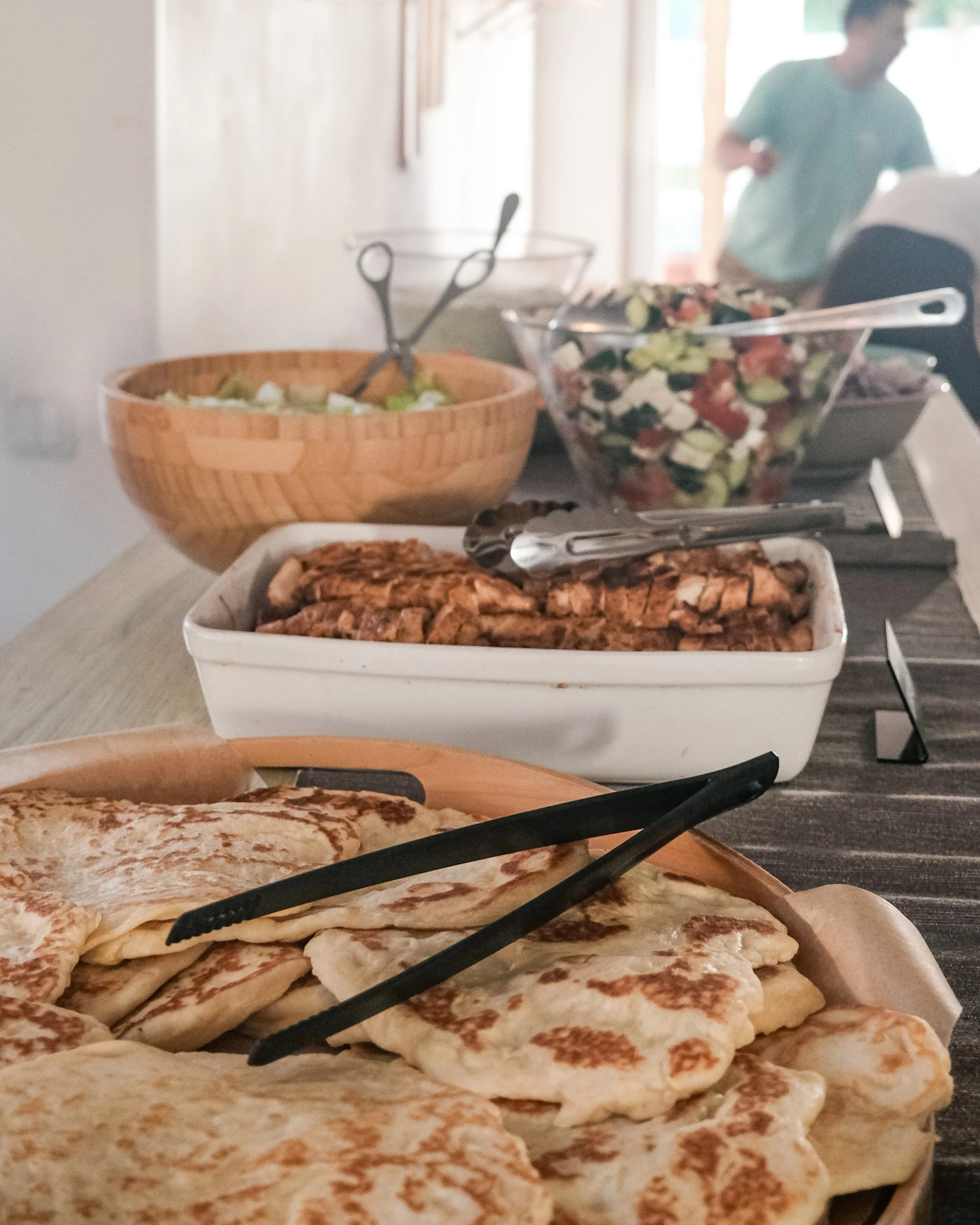 A table with various food items, including a tray of cooked flatbread, a dish of sliced grilled chicken, a bowl of salad, and other dishes in the background, with a person preparing food in the kitchen.