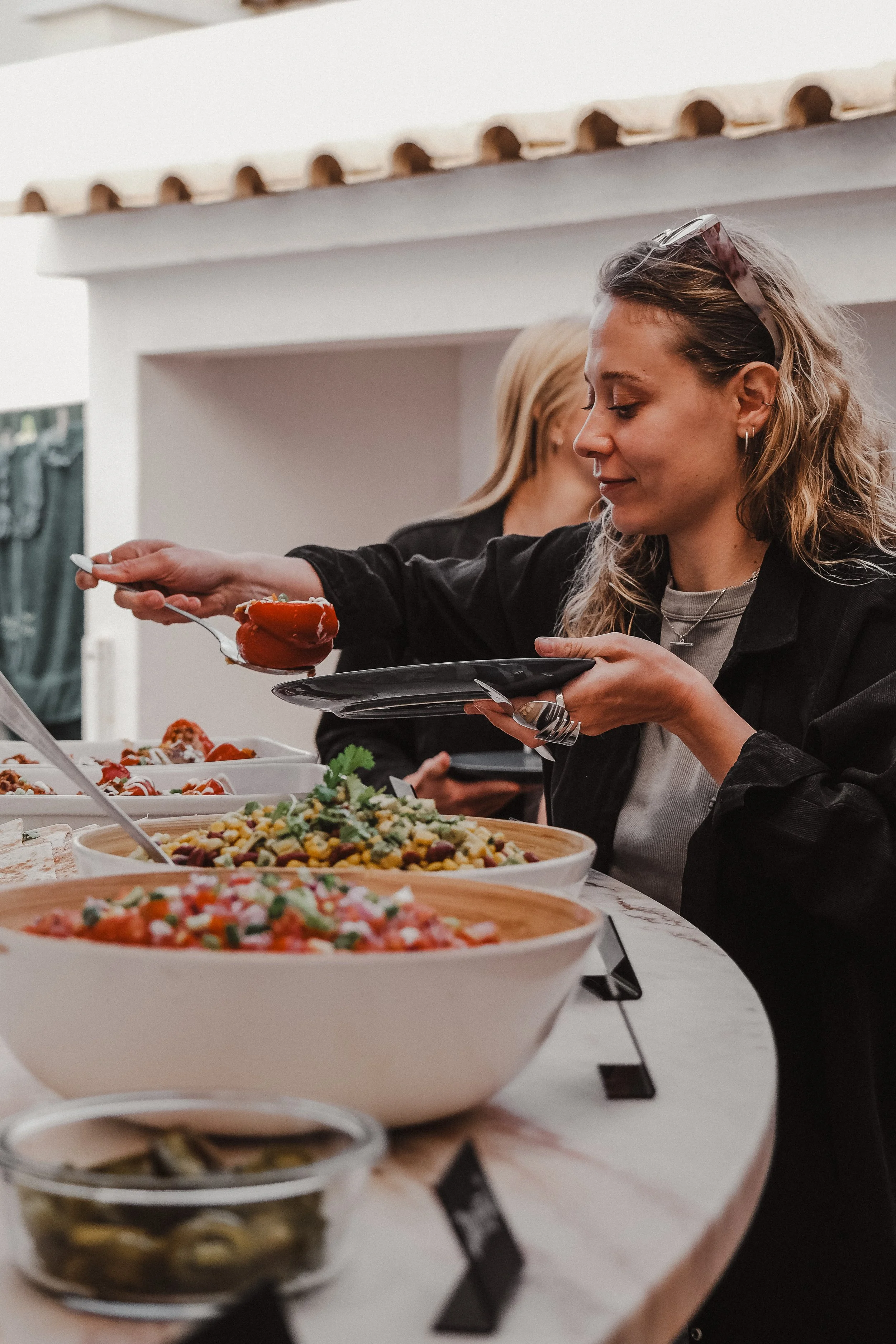Woman serving herself food from a buffet table with various salads and dishes, indoors with a white wall and decorative roof above.