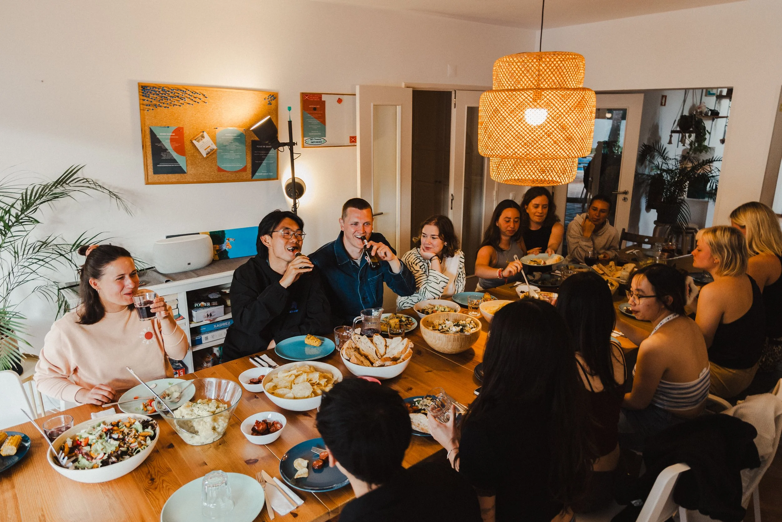 A group of people gathered around a dining table enjoying a meal at a party or celebration in a well-lit room decorated with plants and wall art.