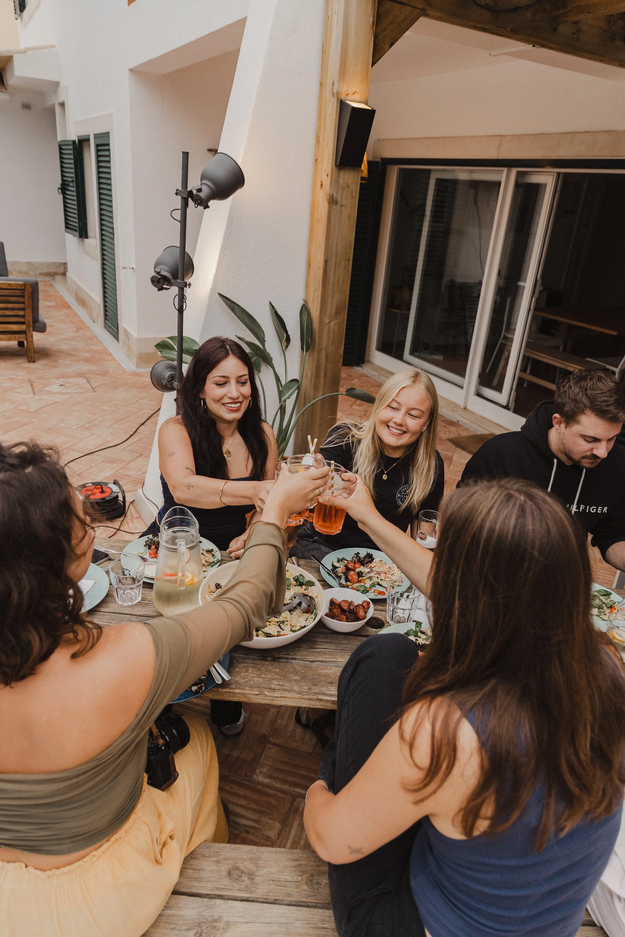 Group of people sitting around a wooden table, toasting with drinks, enjoying a meal at a casual indoor gathering.