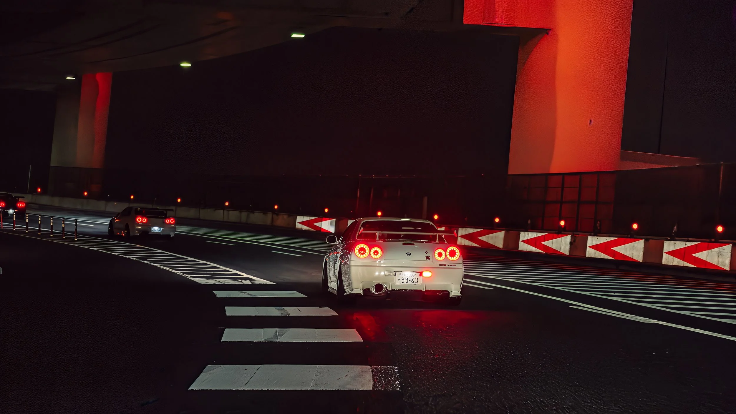 Autos negros y plateados conducen por una carretera nocturna, con paredes y señalización en rojo y blanco, en un túnel con iluminación tenue.