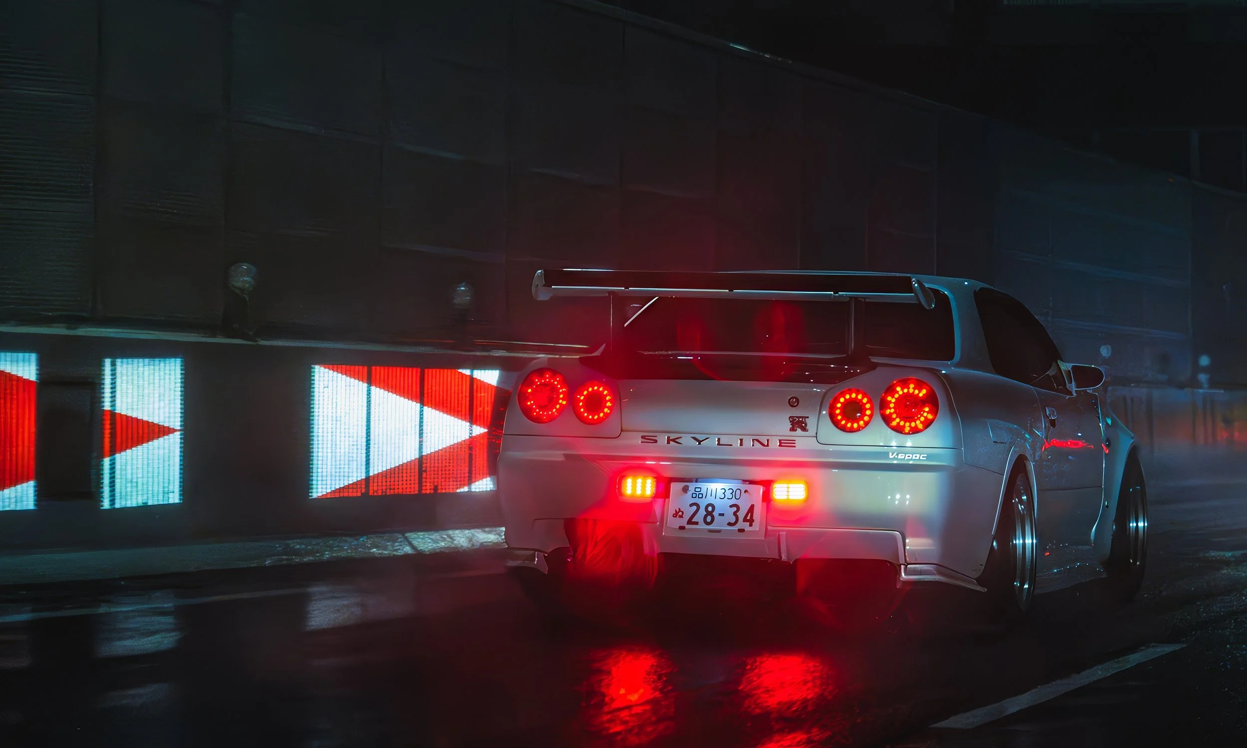 Autos deportivos Nissan Skyline en la noche con luces de freno y una señal luminosa con flechas en la vía, en un entorno oscuro y húmedo.