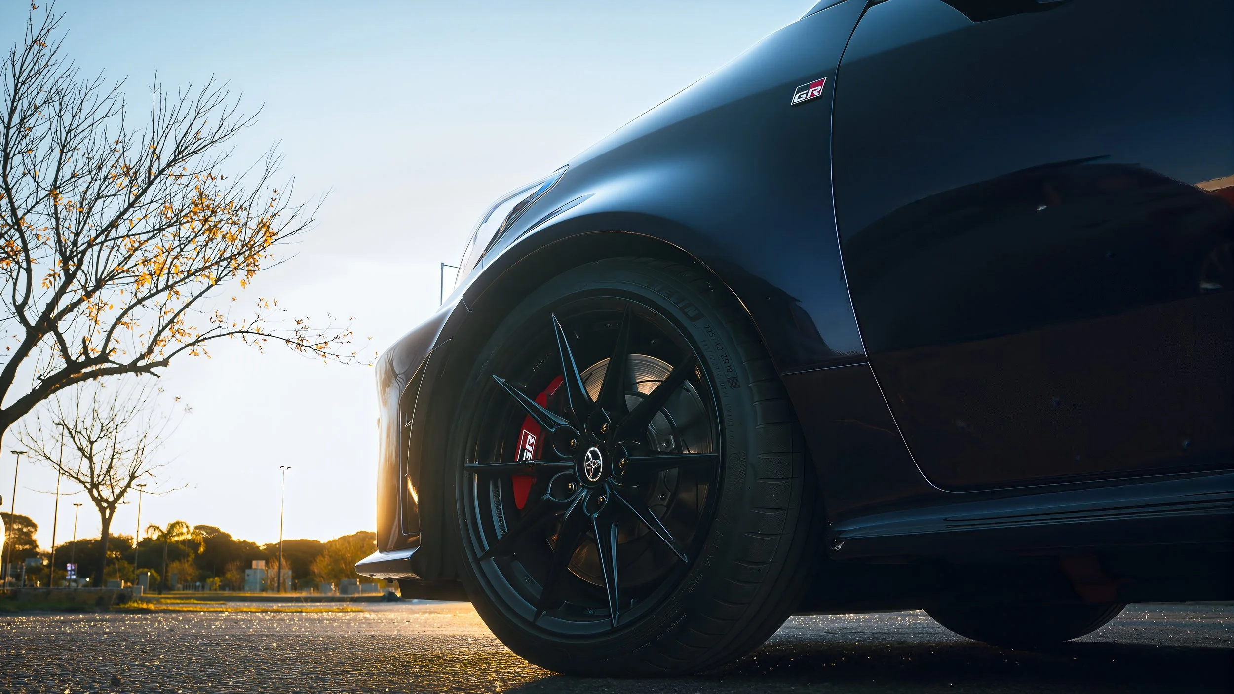 Vista de un auto deportivo negro con llantas de carreras y calipers rojos, al atardecer en un estacionamiento con árboles y cielo claro.