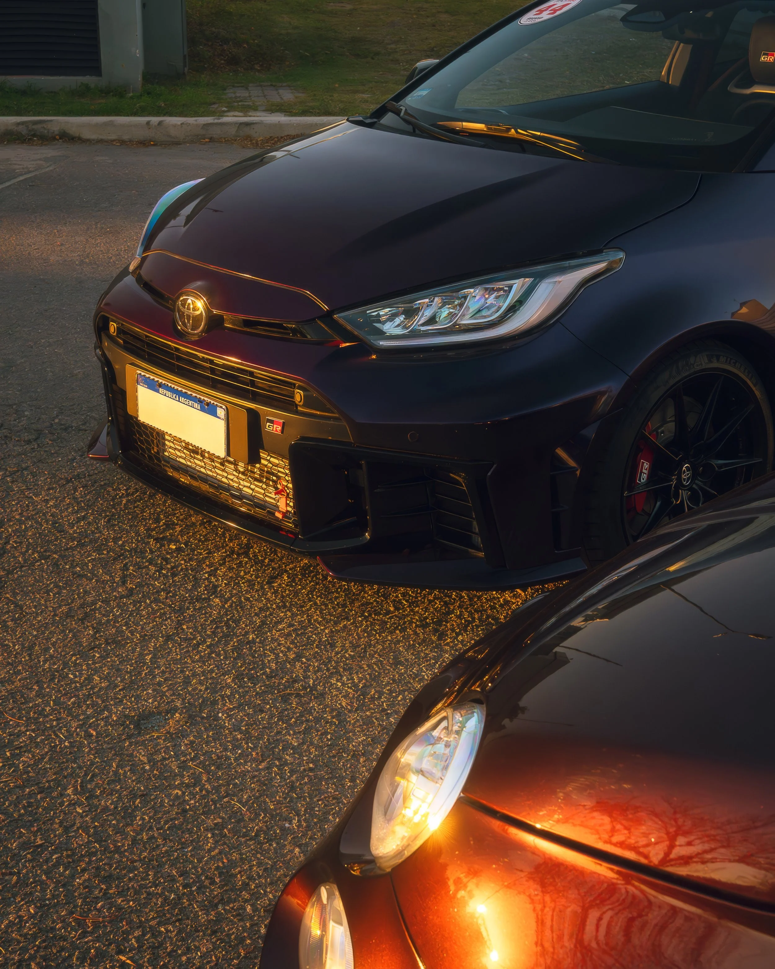 Primer plano de un coche oscuro y un coche rojo, con las luces delanteras encendidas, estacionados en la calle en la tarde, reflejando el entorno en sus superficies.