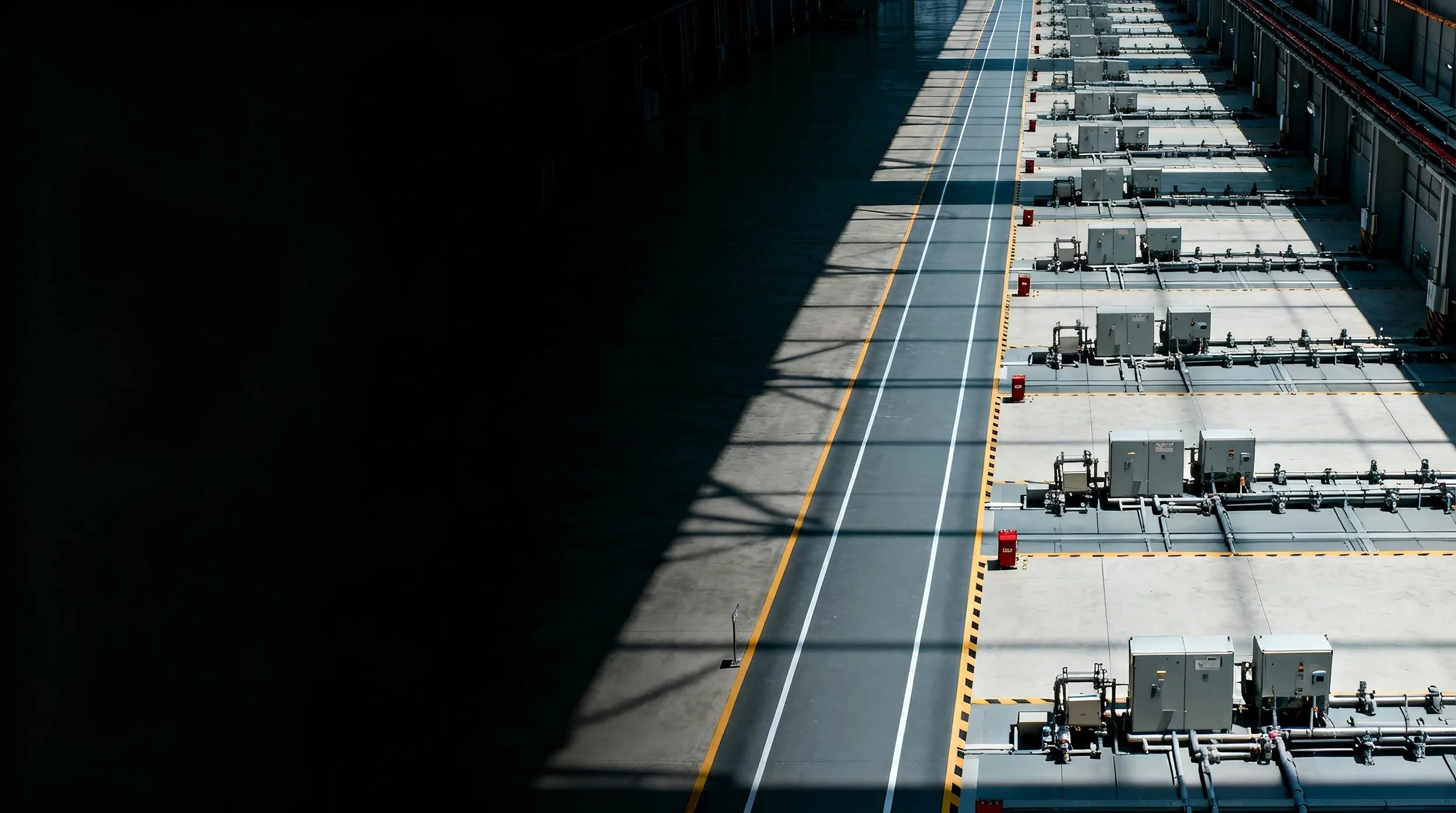 Empty parking lot with industrial equipment and pipelines, partially in shadow and partially in sunlight.