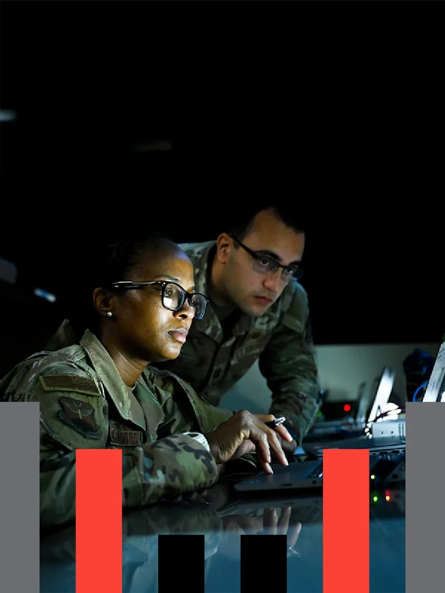 Two military personnel working on computers in a dark room, focused on screens. One woman and one man, both in camouflage uniforms and glasses.