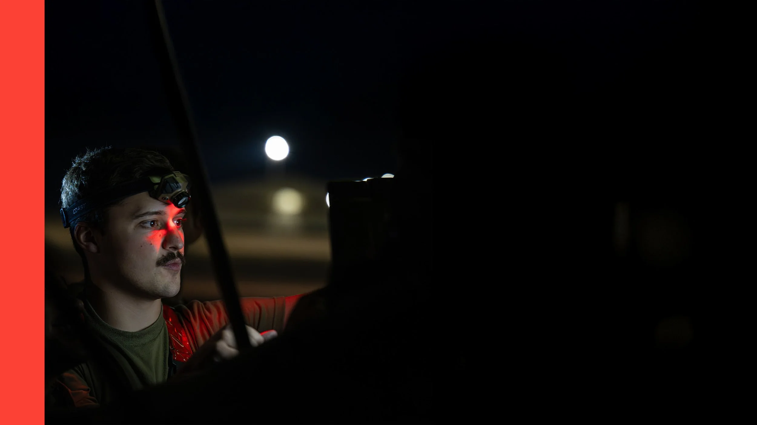A man with dark hair and a mustache wearing a headlamp, sitting in a vehicle at night, with a bright moon in the sky outside.