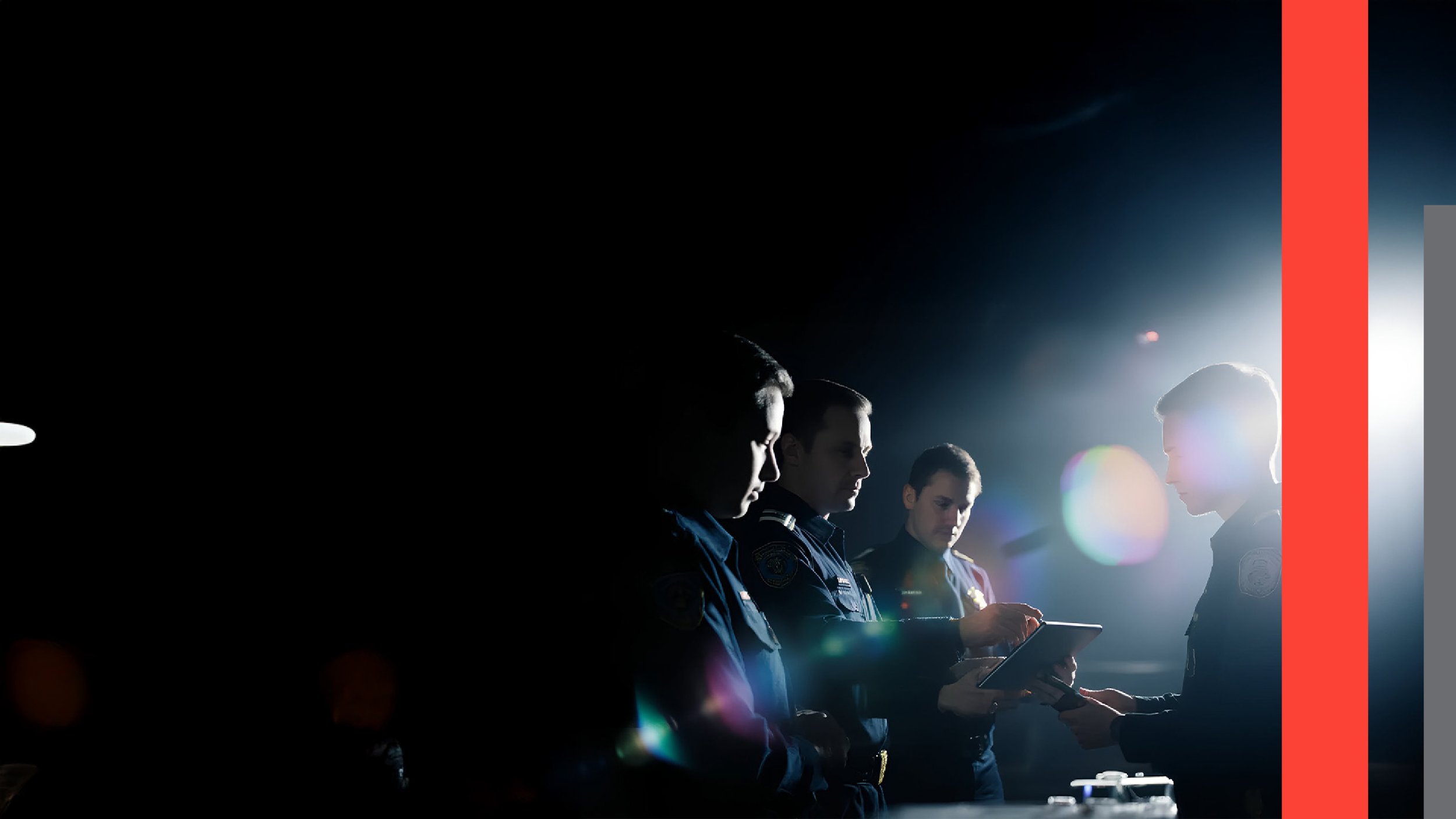 Four police officers, two men and two women, standing in a row in police uniforms, with one woman handing an object to the other woman, in a dark setting illuminated by bright light from behind.