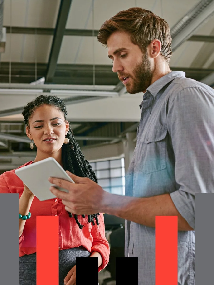 Two people, a woman with curly hair and a man with a beard, are looking at a tablet together in an office setting.