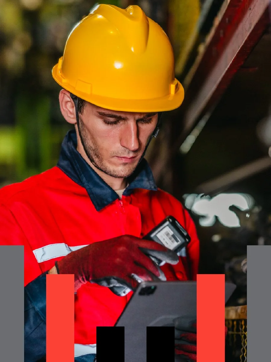 A male worker wearing a yellow safety helmet and red work jacket, focused on a device in his hand, standing outdoors at night.