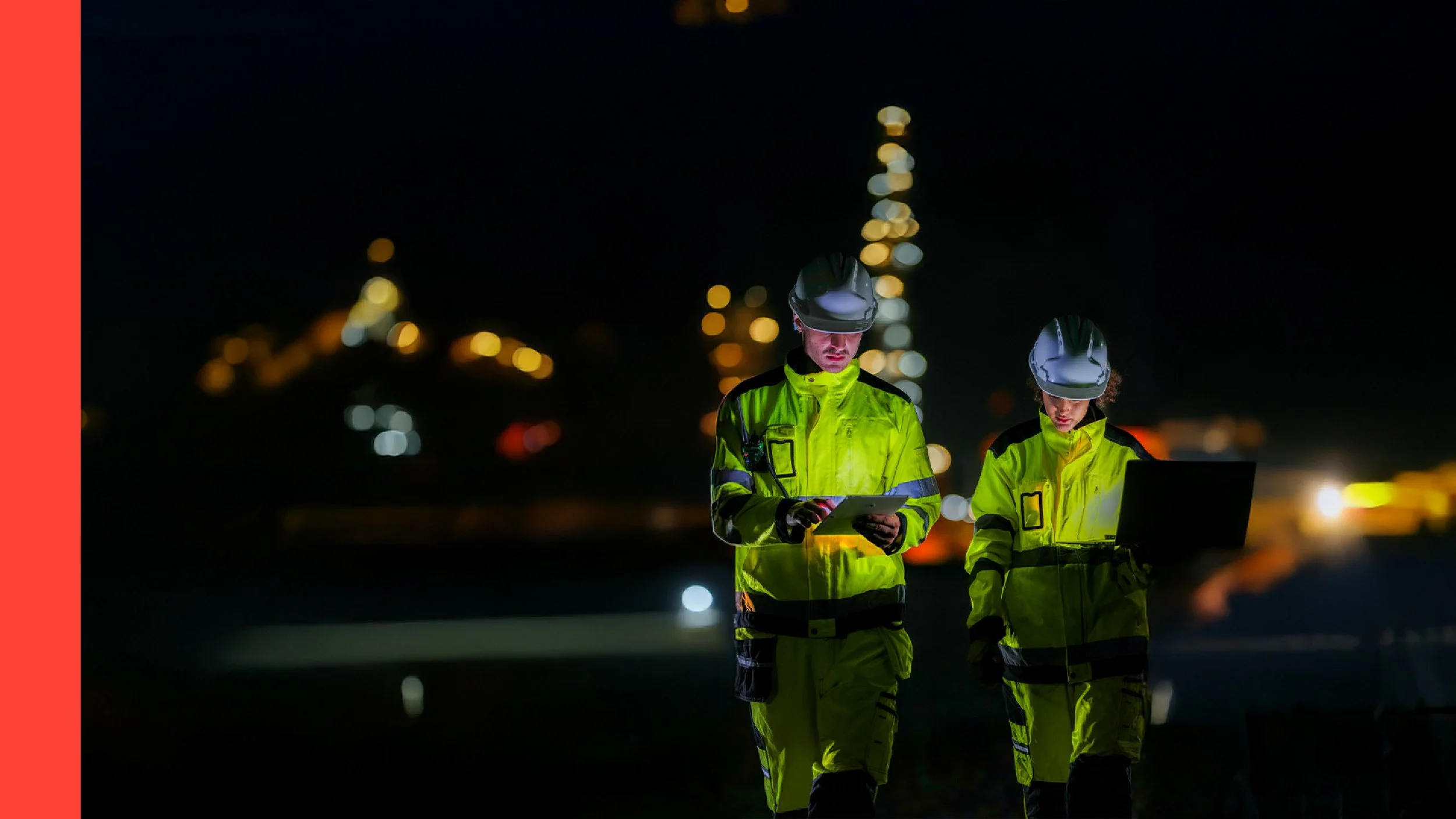 Two emergency responders in high-visibility yellow jackets and helmets walk at night, checking electronic devices, with city lights blurred in the background.