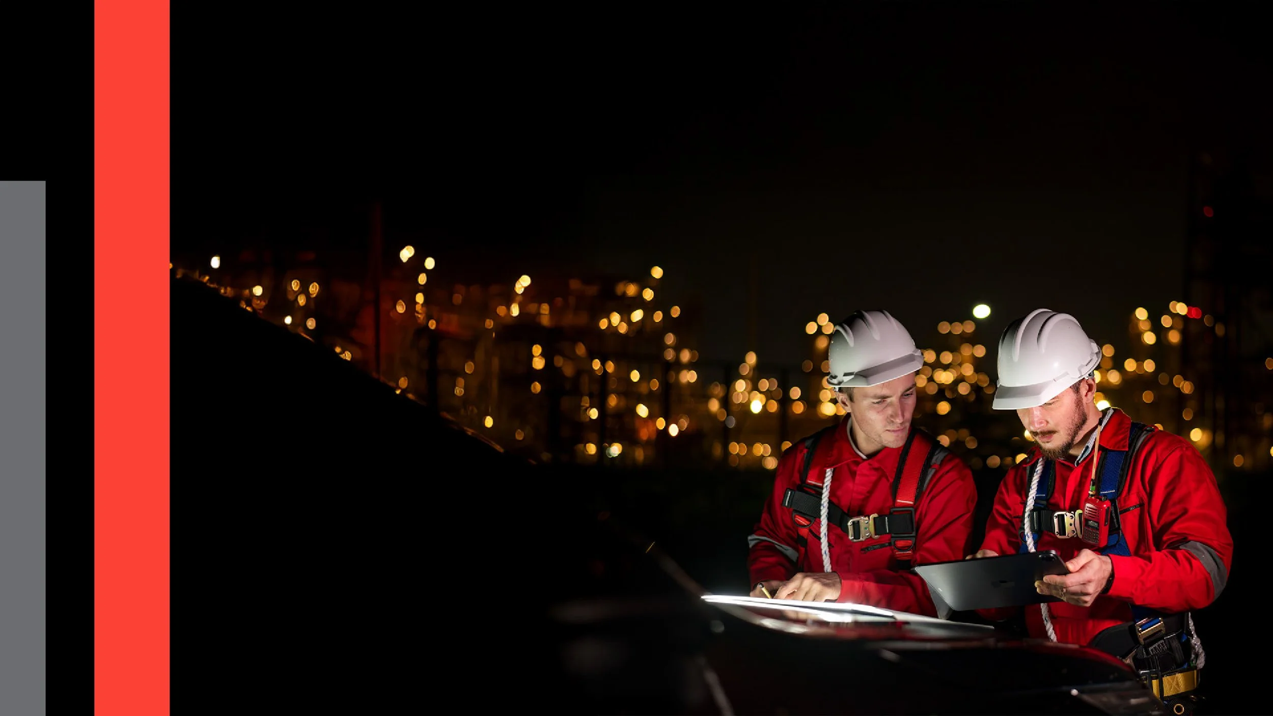 Two construction workers in red uniforms and white helmets examining documents and using a tablet at night, with a blurry city skyline in the background.