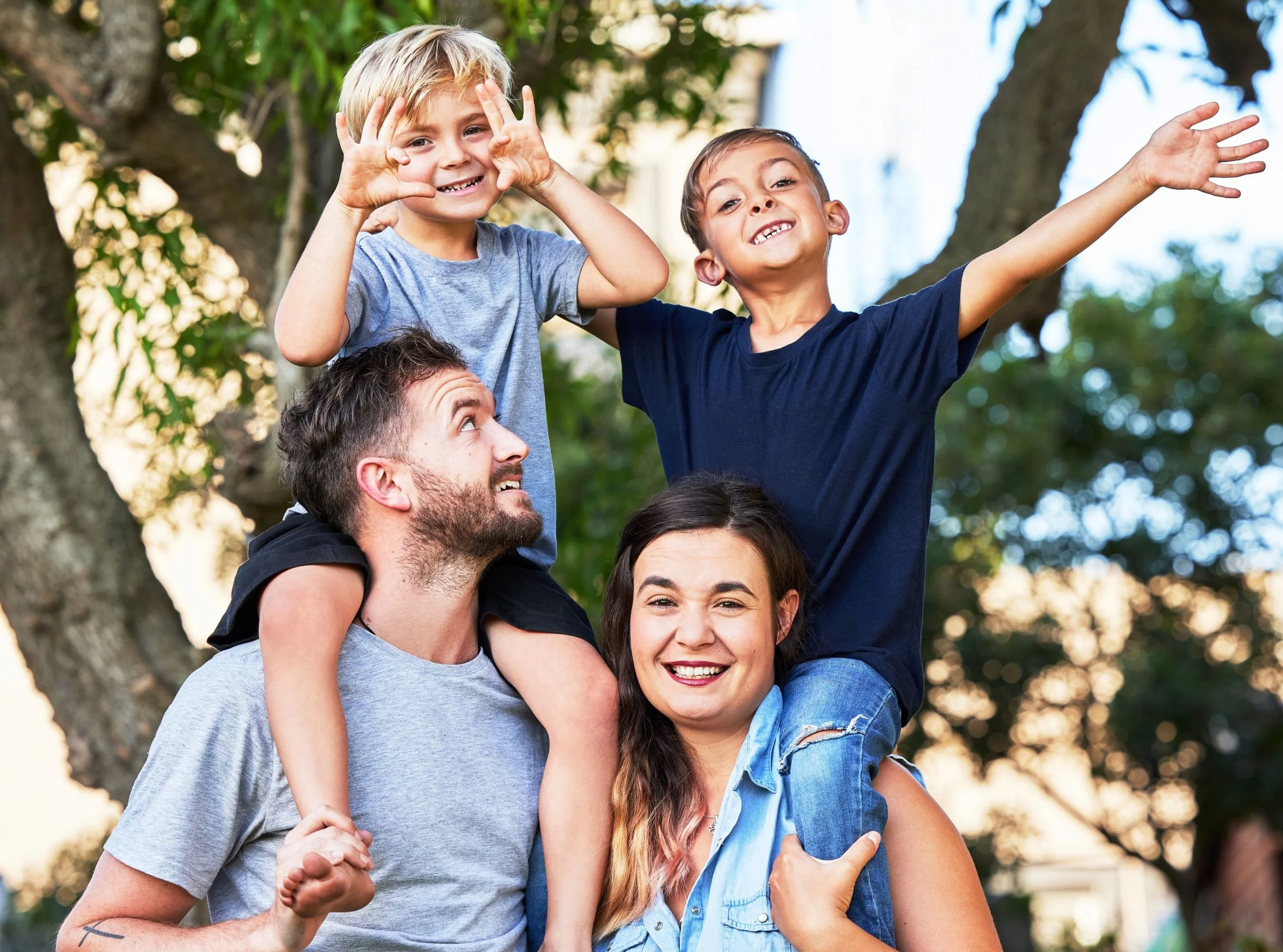A happy family of four, with two children sitting on their parents' shoulders, smiling and enjoying time outdoors in a park with trees.